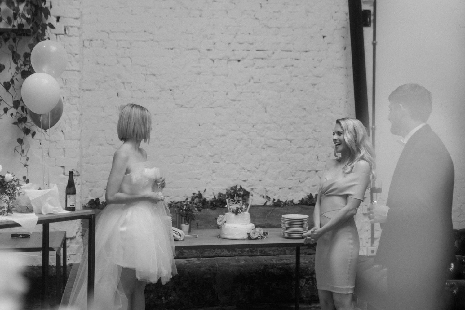 A celebration scene with guests gathered around a dessert table against white brick walls.