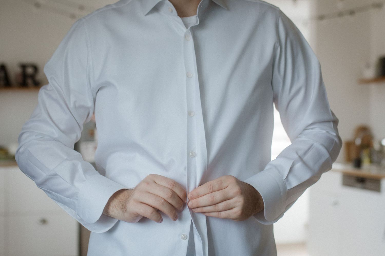 Person buttoning up a crisp light blue dress shirt in a bright kitchen setting.