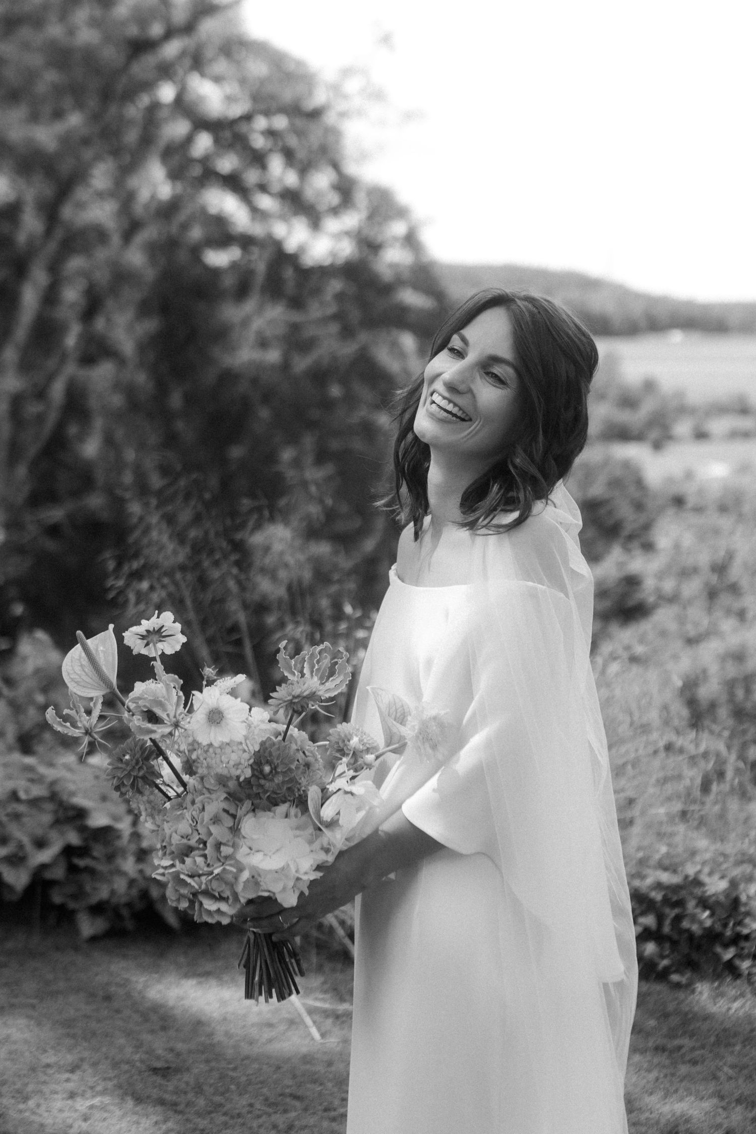 Black and white portrait of a joyful bride holding a floral bouquet in an outdoor setting.