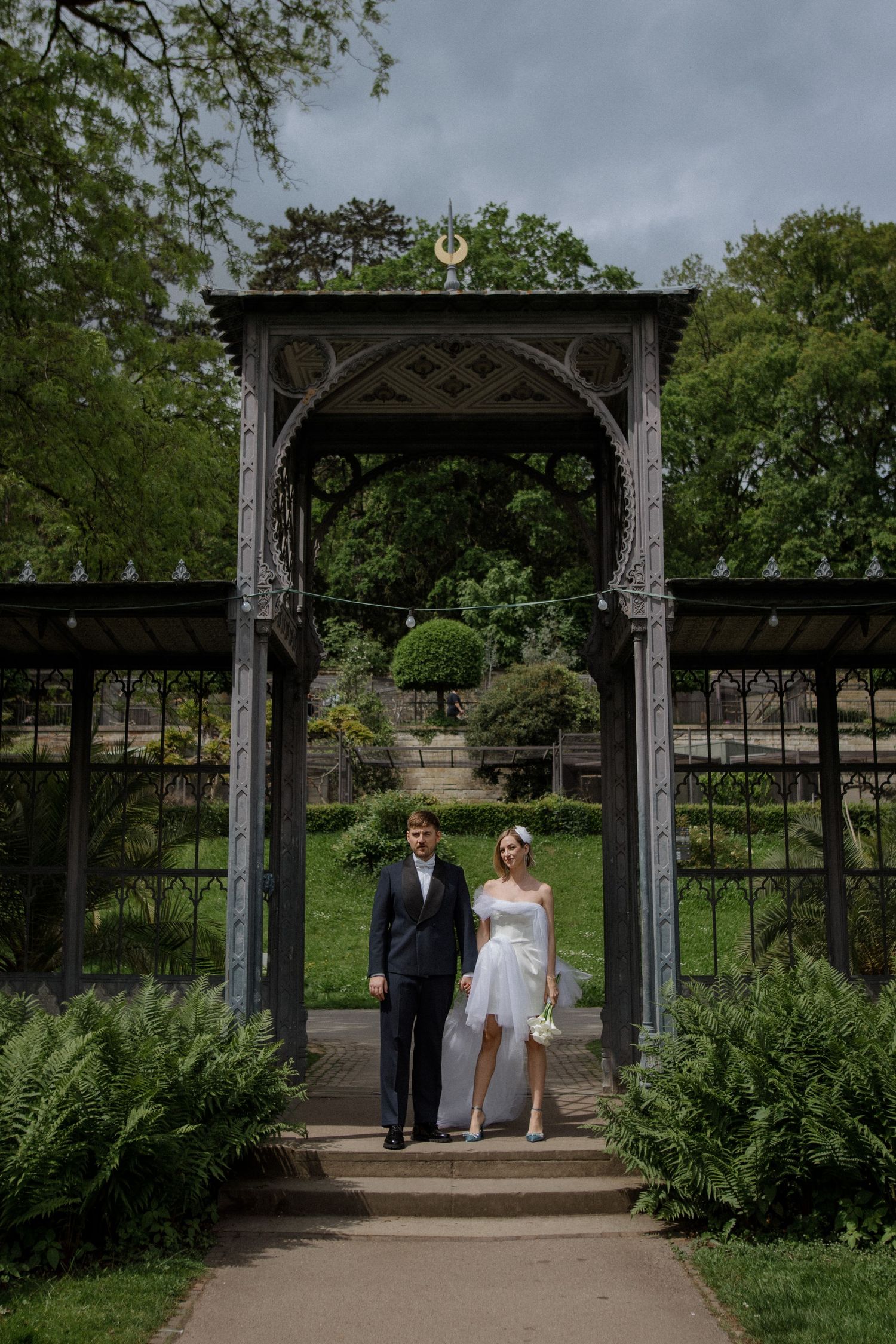 Wedding couple standing under an ornate Victorian garden archway surrounded by greenery.