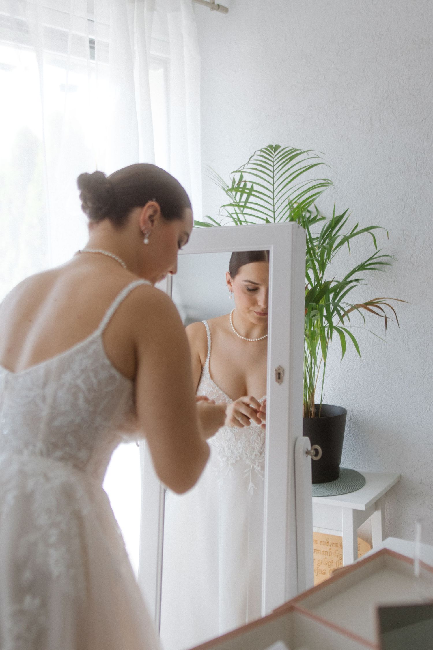 Wedding preparation scene with white lace dress and mirror reflection near a potted palm plant.