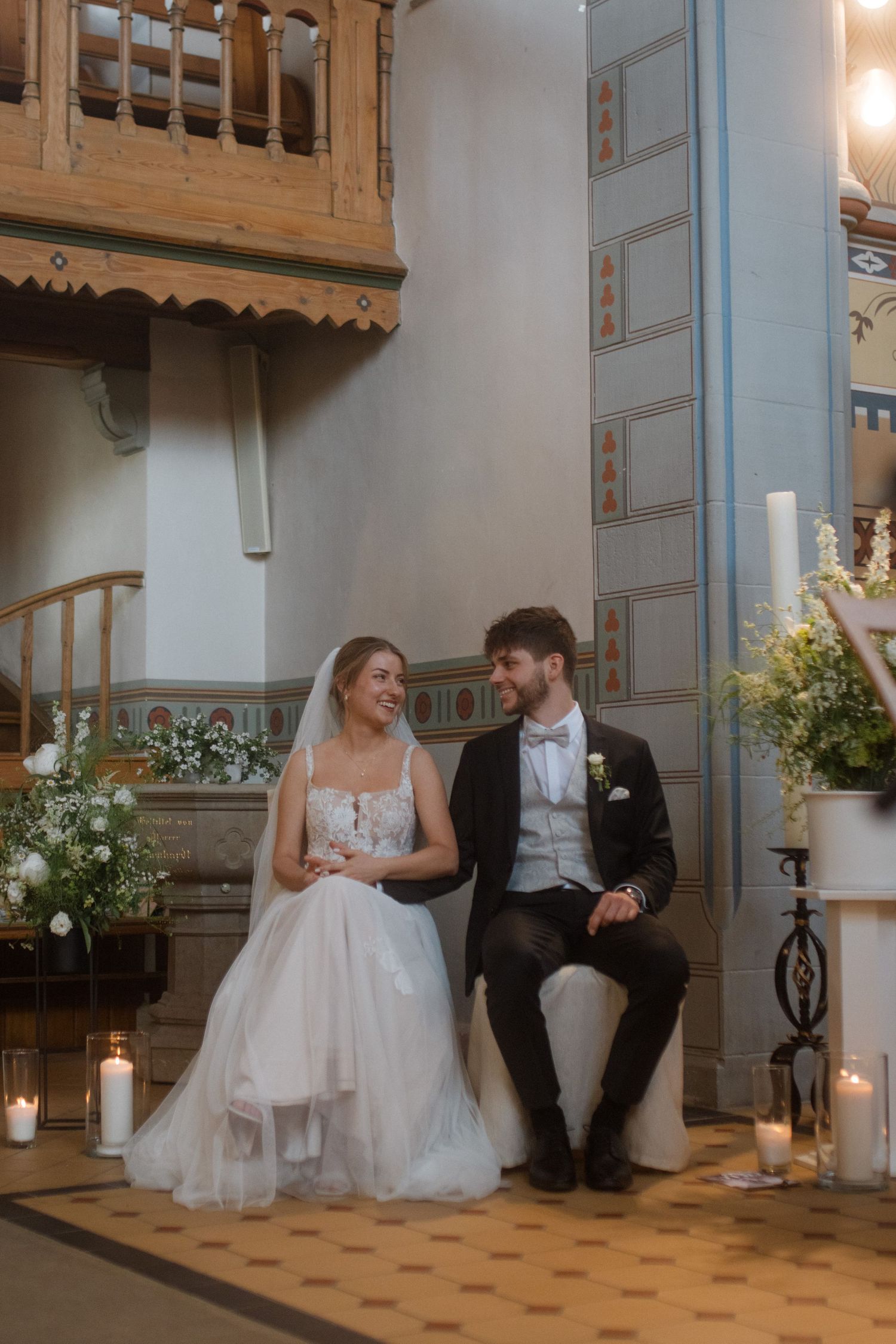 A newlywed couple sits surrounded by candles and floral arrangements in a historic church setting.