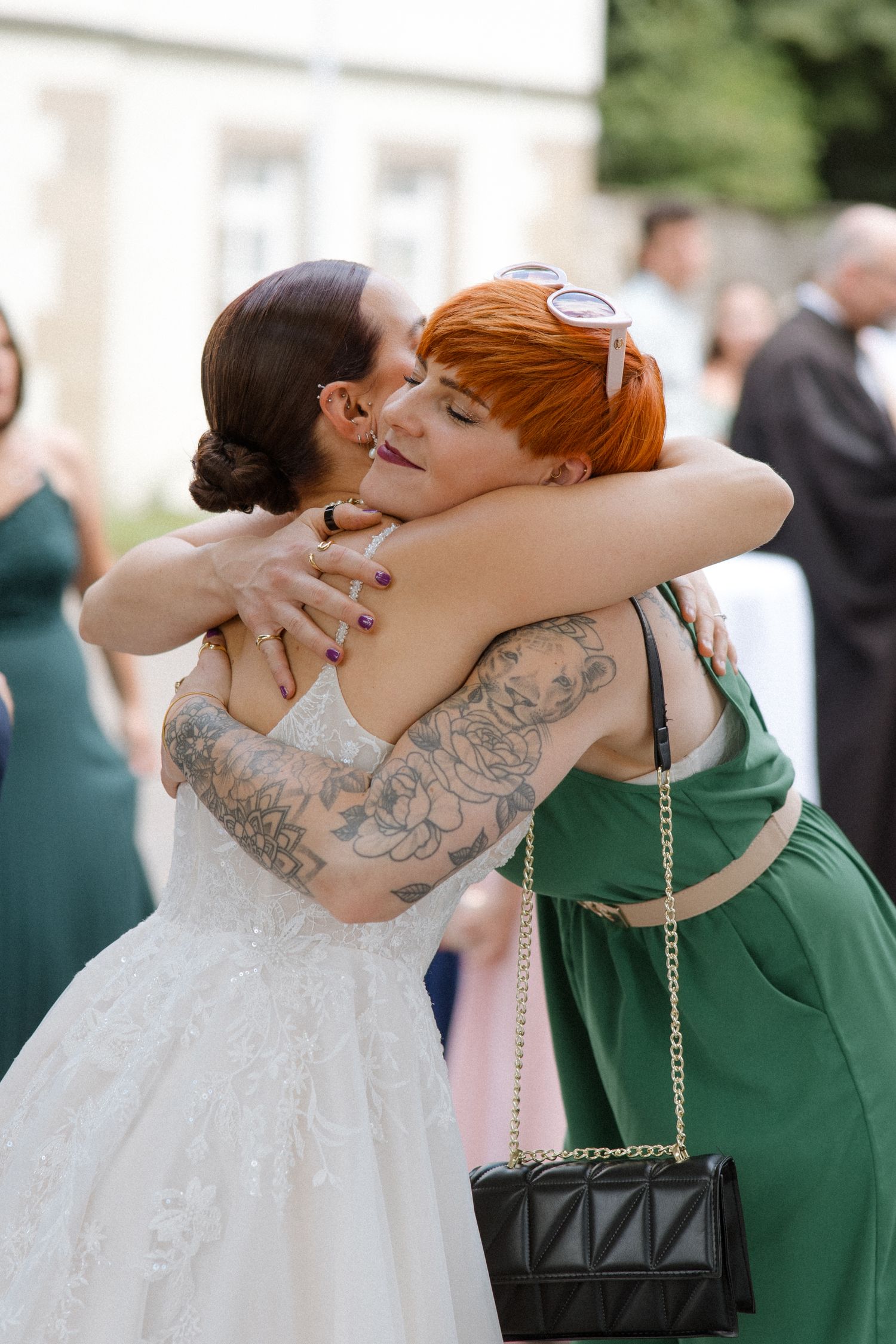 A bride in a lace dress hugs a guest wearing a green dress and displaying colorful tattoos.