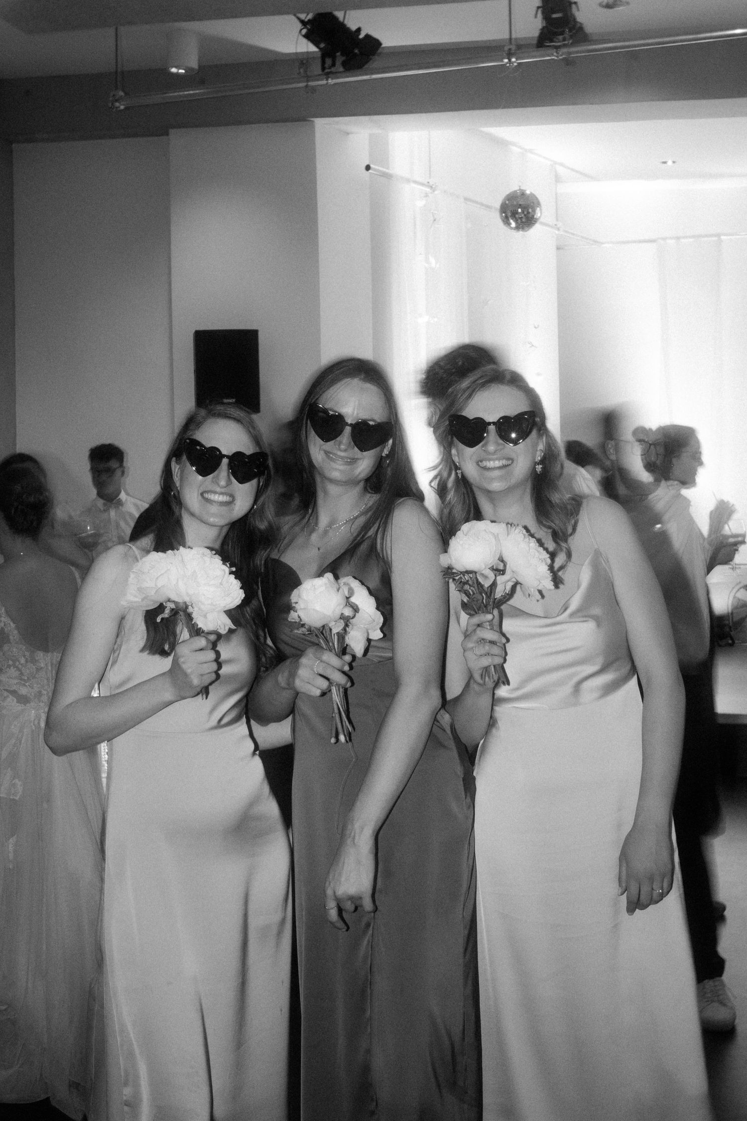 Three bridesmaids in formal dresses pose with flowers while wearing sunglasses.
