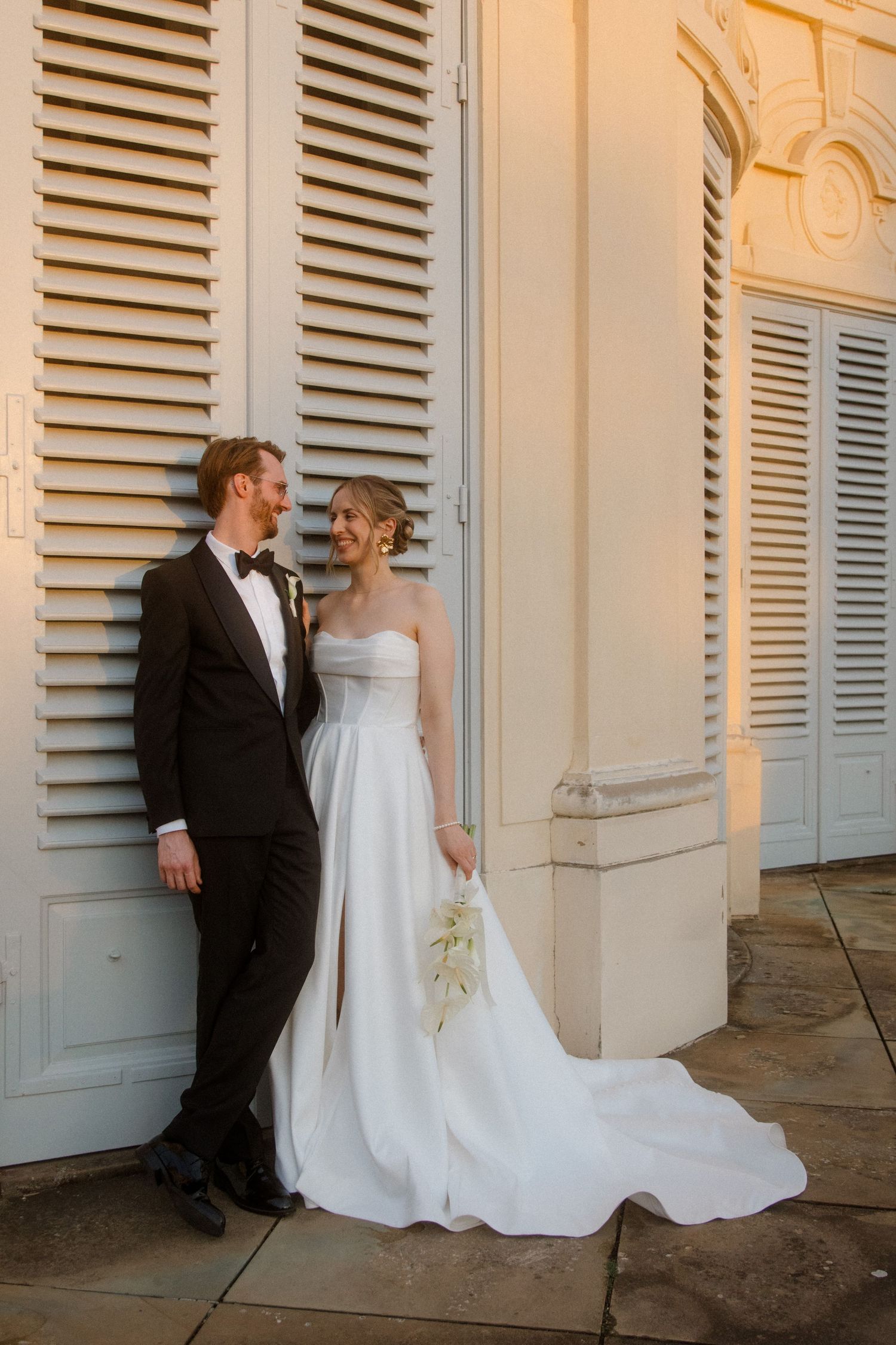 Wedding couple posing against a cream-colored wall with blue shutters at sunset.
