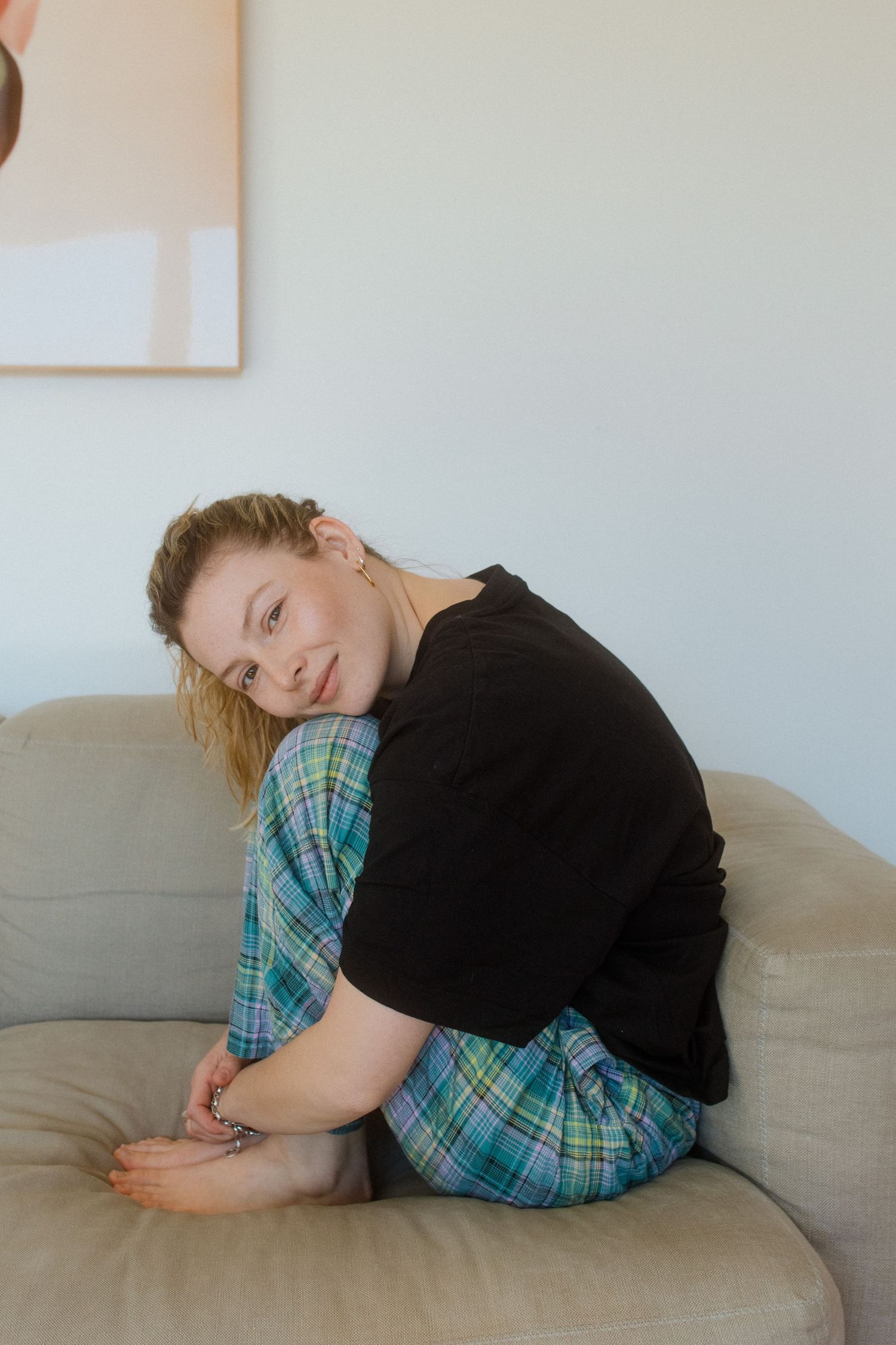 A person wearing plaid and black lies curled up in a fetal position on a beige couch in a minimalist setting.