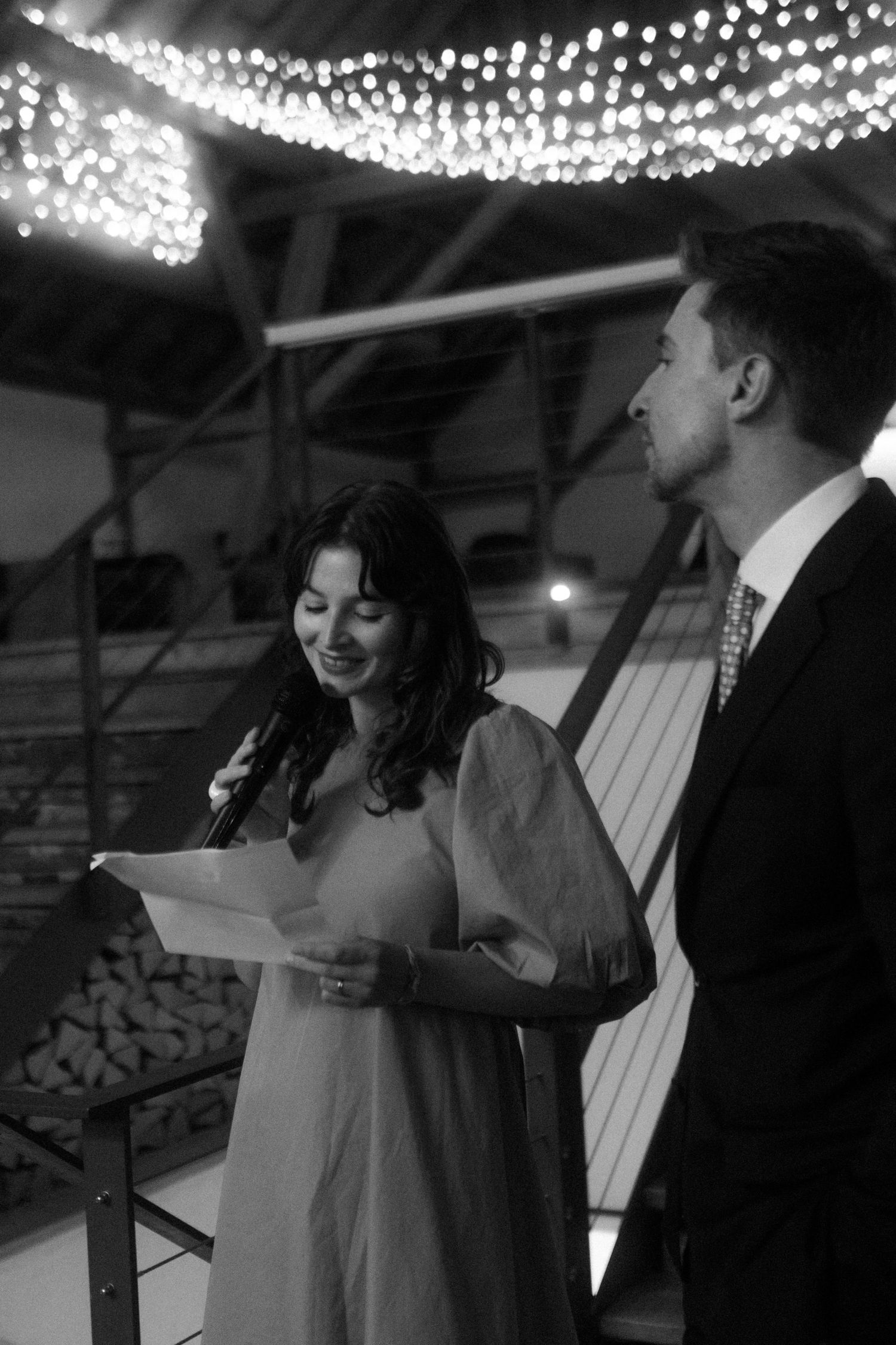 Two people speaking at an event under twinkling lights in rustic venue.