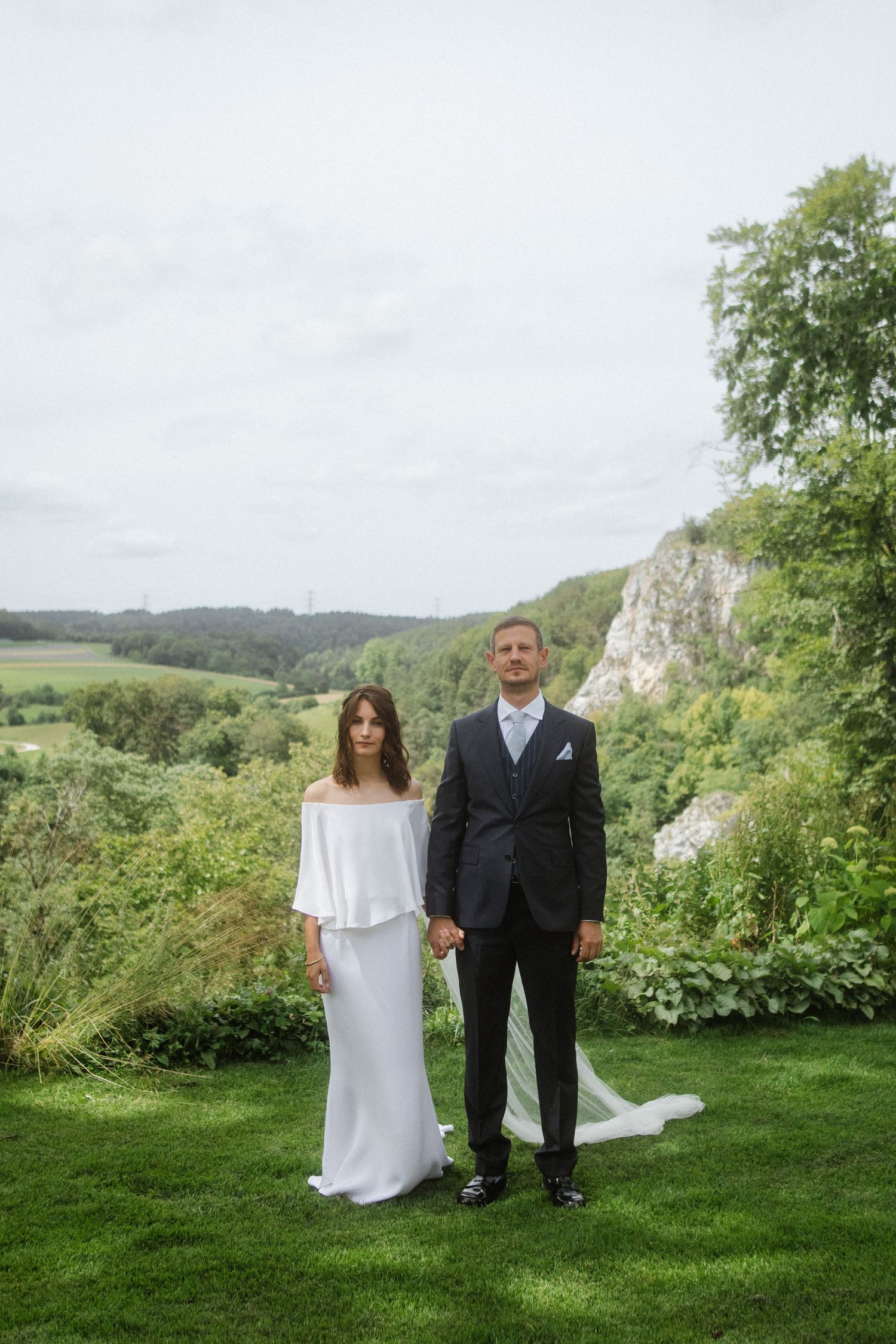 A wedding portrait of a couple standing together in an elegant garden with dramatic cliffs behind them.