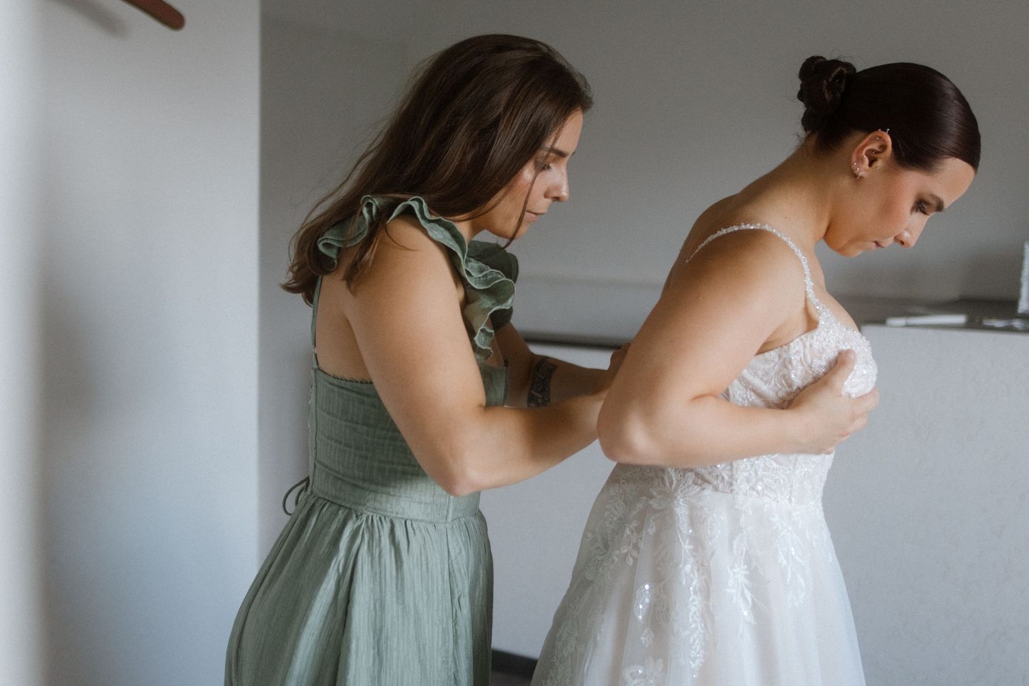 Helper in sage green dress assisting with wedding gown fitting.