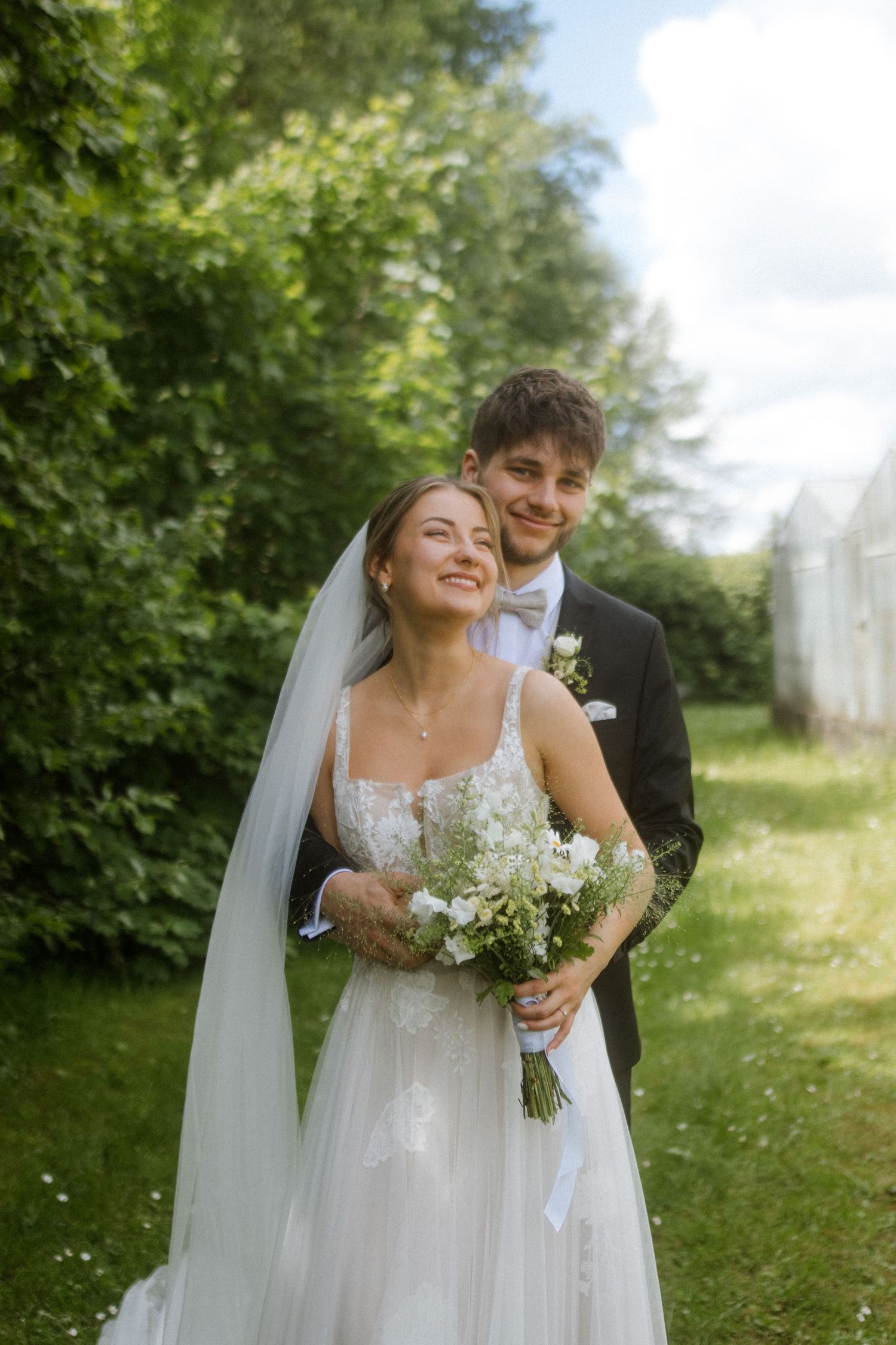 A joyful wedding moment captured in a garden setting with white greenhouses visible in the background.