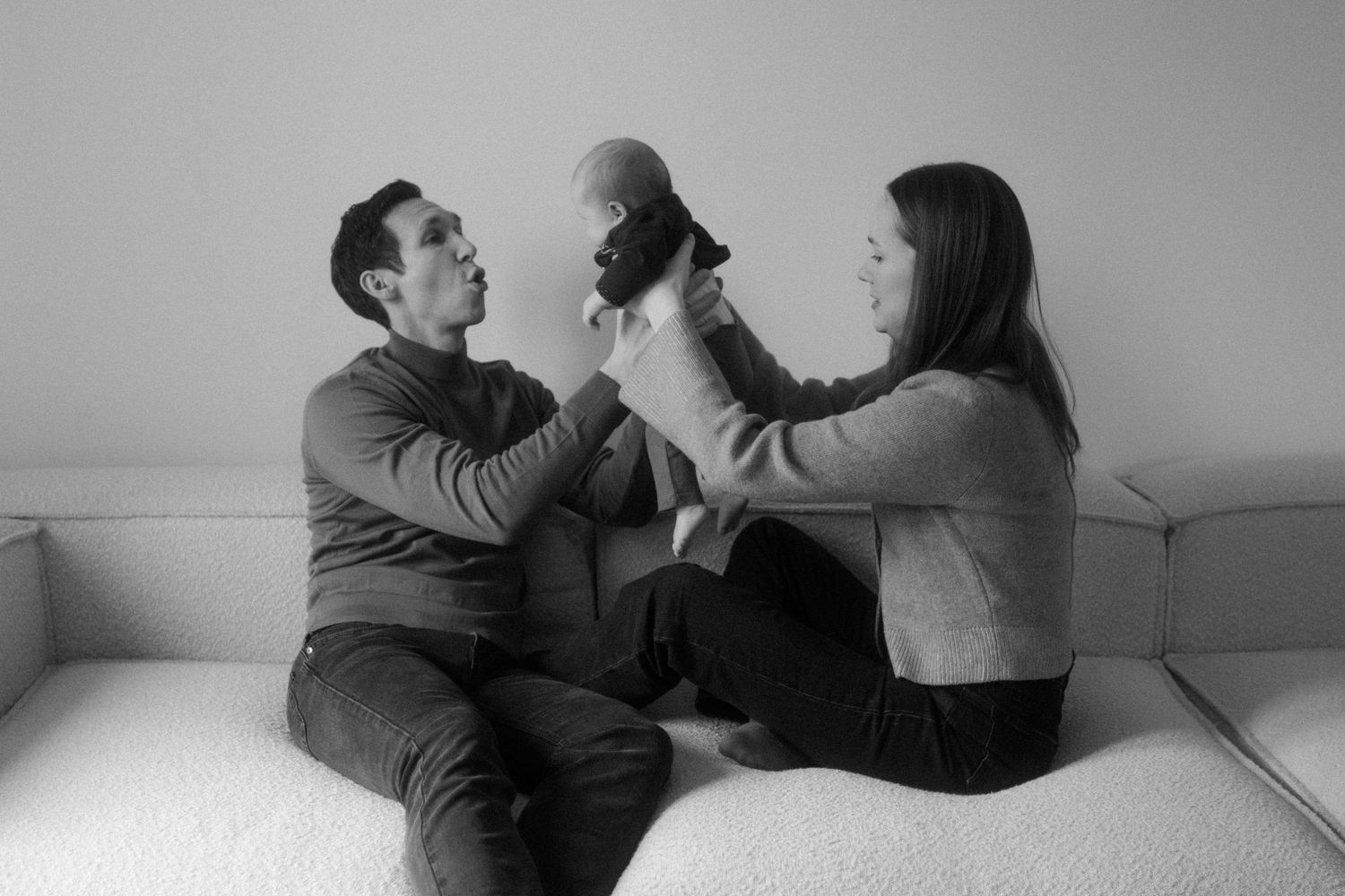 A couple sits on a white couch playing with their black cat in a black and white photograph.
