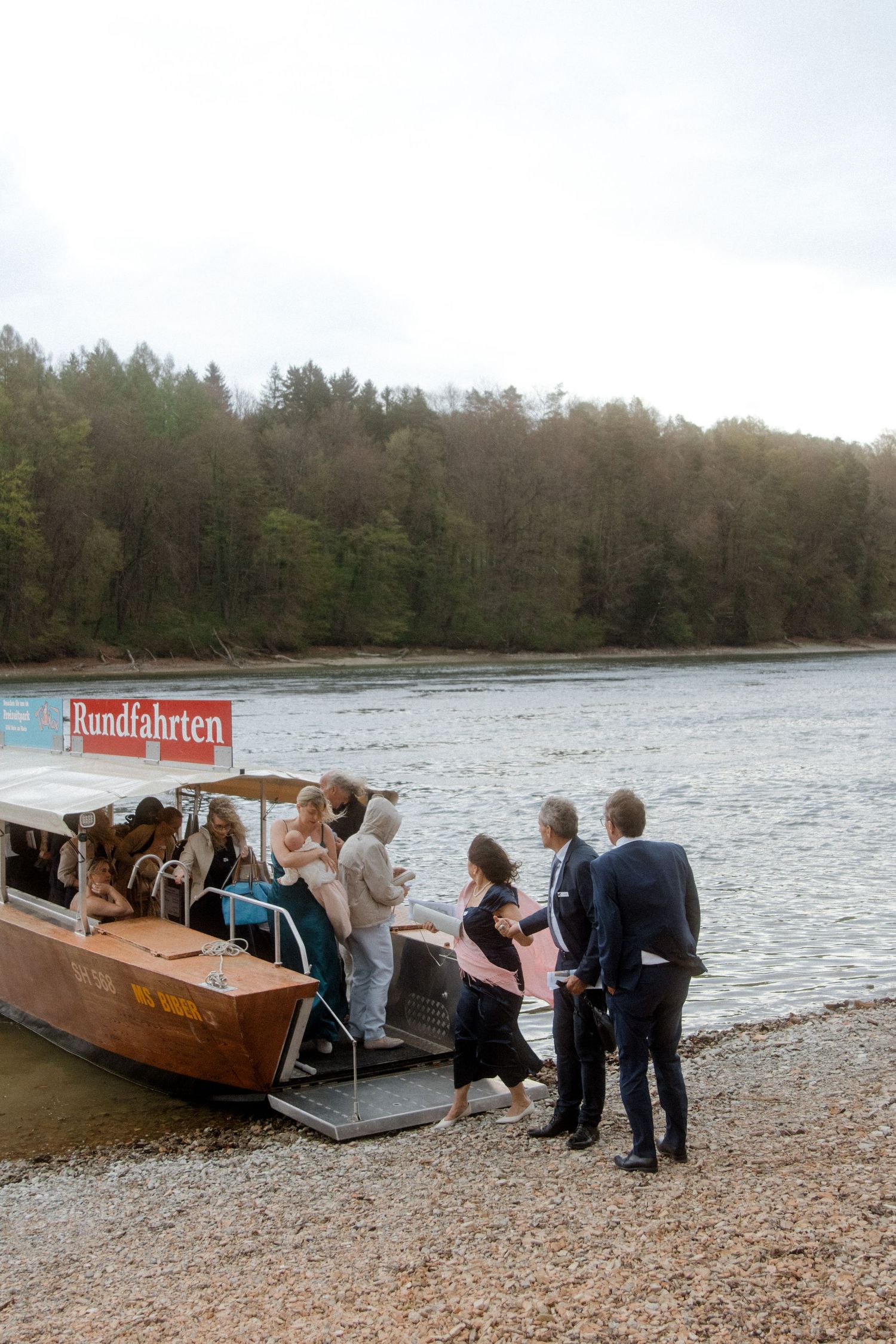 Wedding guests board a wooden Rundfahrten tour boat on a calm river surrounded by lush green forest.