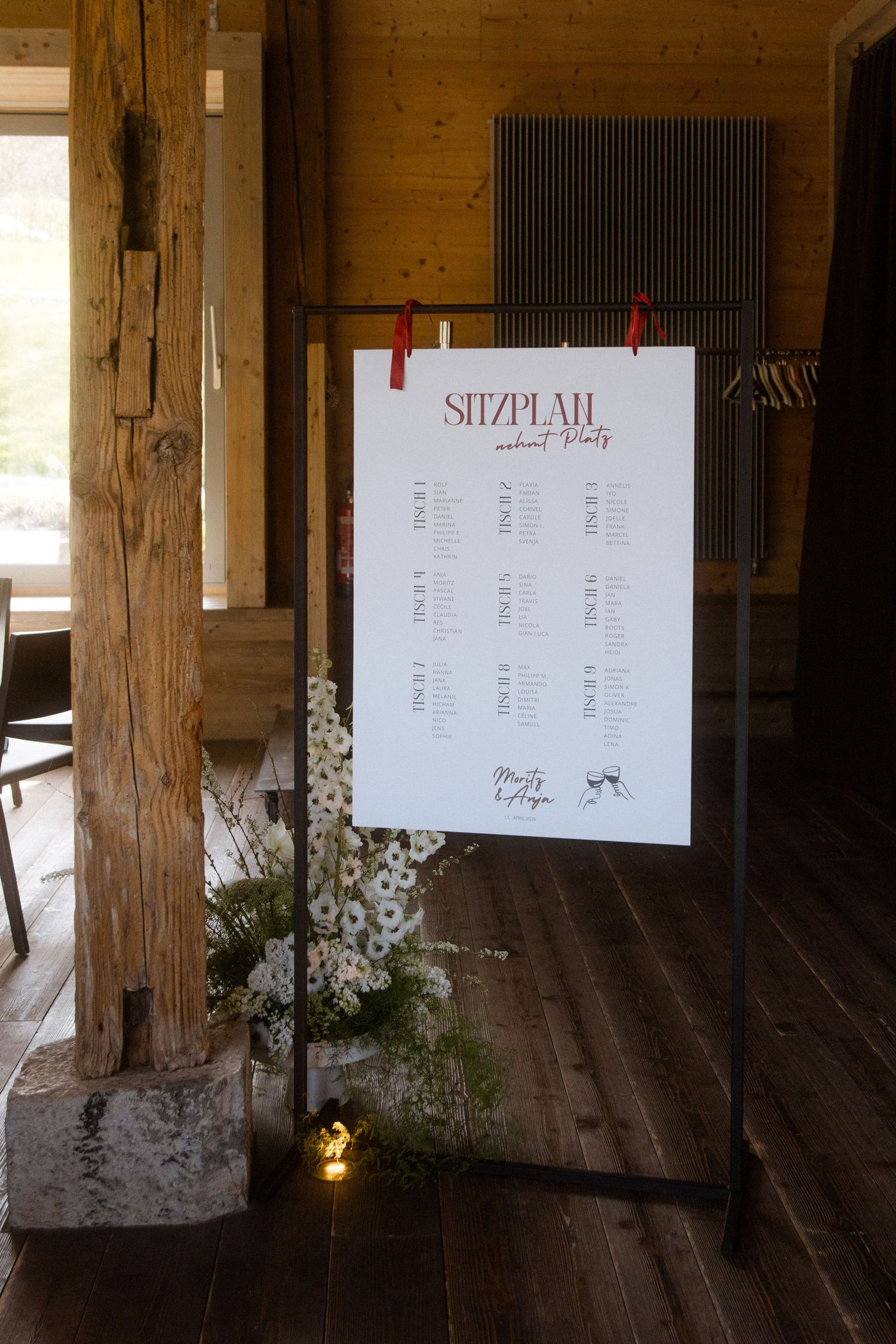 Elegant wedding seating chart sign on a metal stand with red clips, decorated with white flowers in a rustic barn venue.
