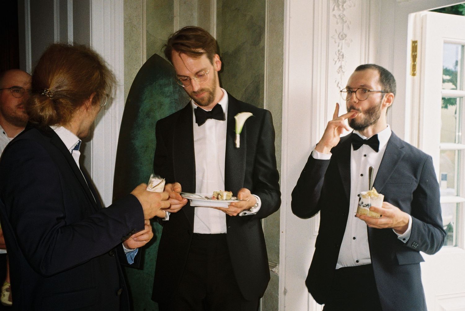 Groomsmen in black tuxedos help each other get ready before a formal wedding ceremony.