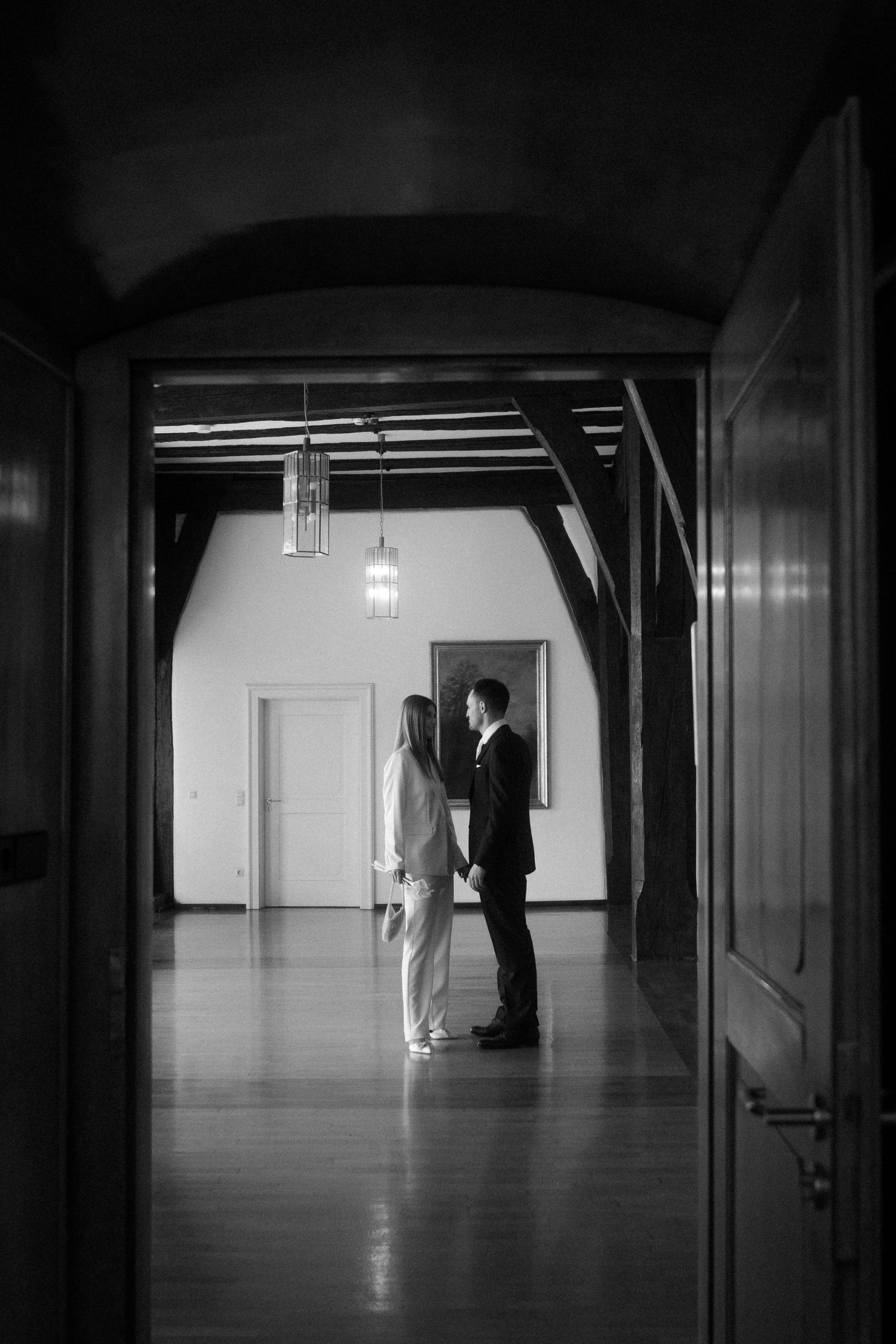 Black and white photo of a couple holding hands in an elegant hall with wooden beams and pendant lights.