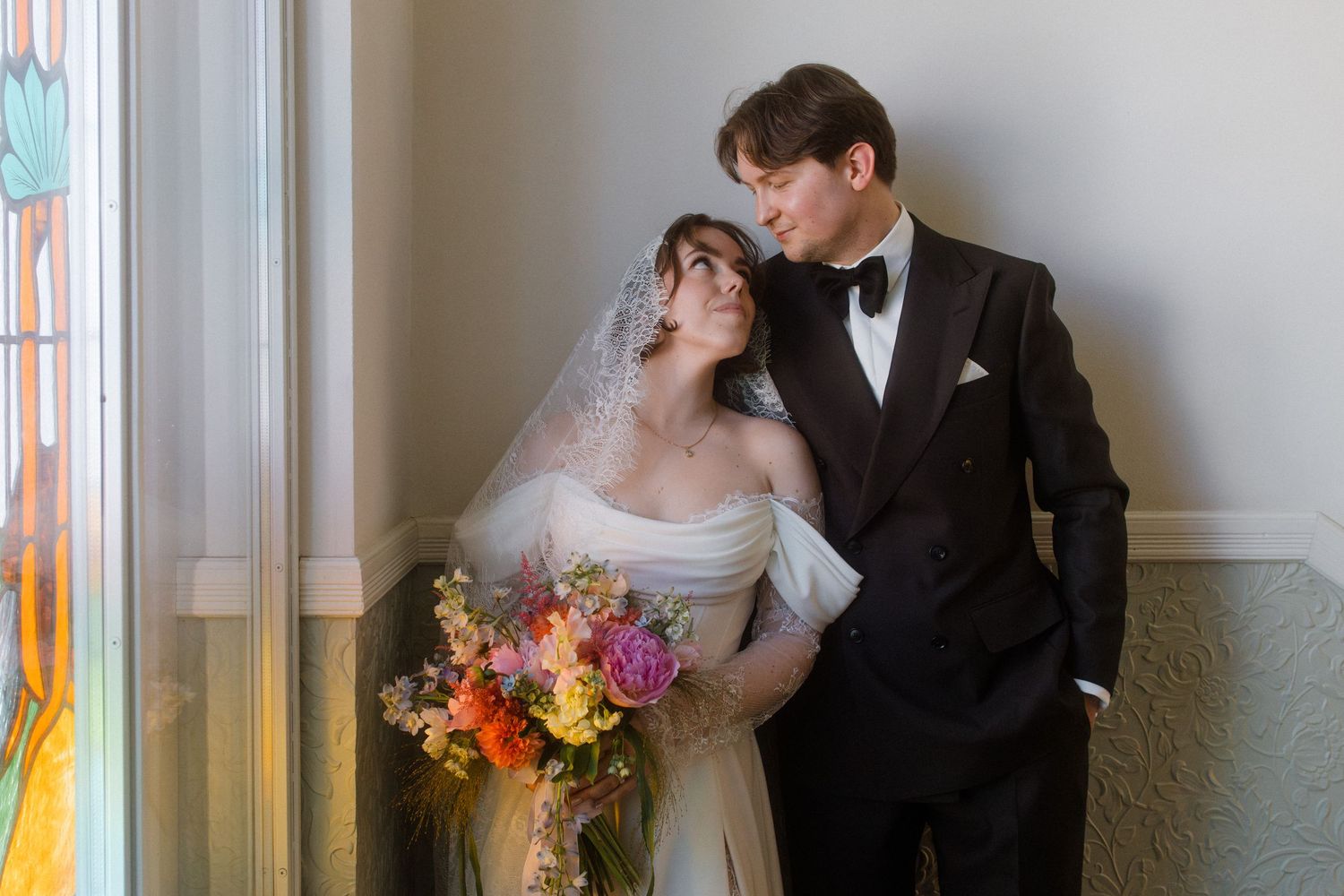 Bride in off-shoulder gown with veil holds colorful bouquet beside groom in black tuxedo near window.
