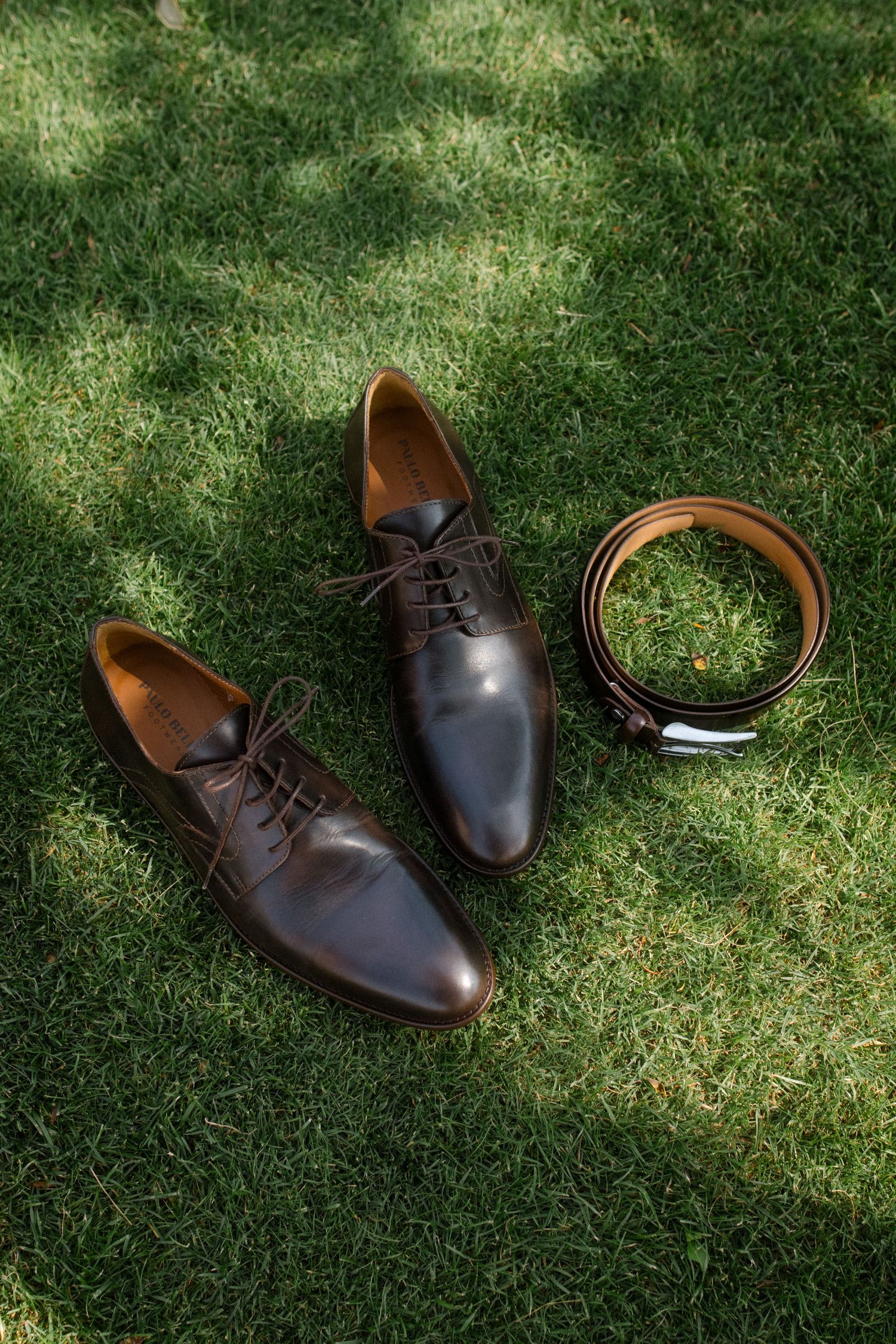 Brown leather dress shoes and belt arranged on green grass in sunlight.