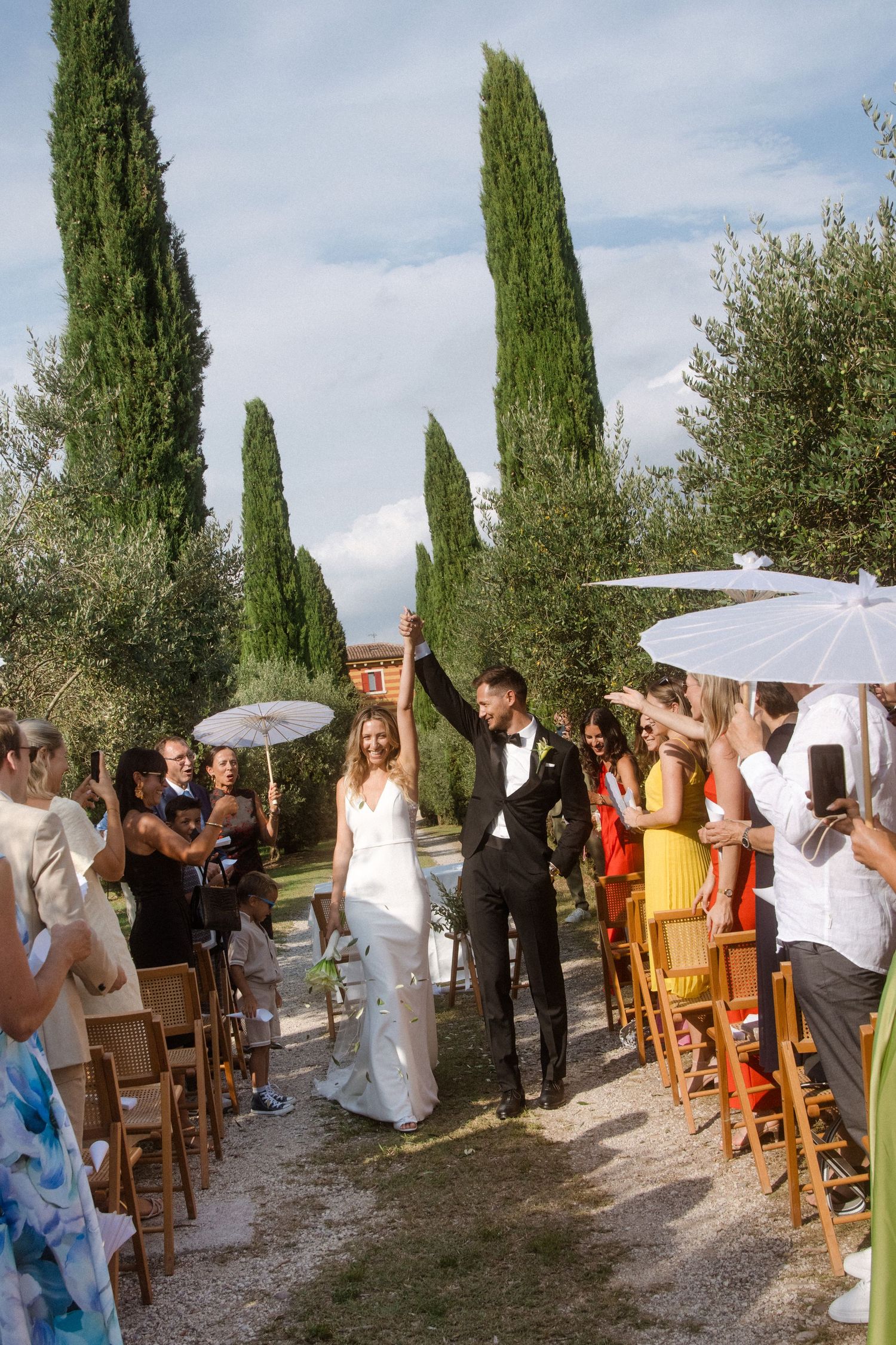 Wedding recessional along cypress-lined path with guests holding parasols.