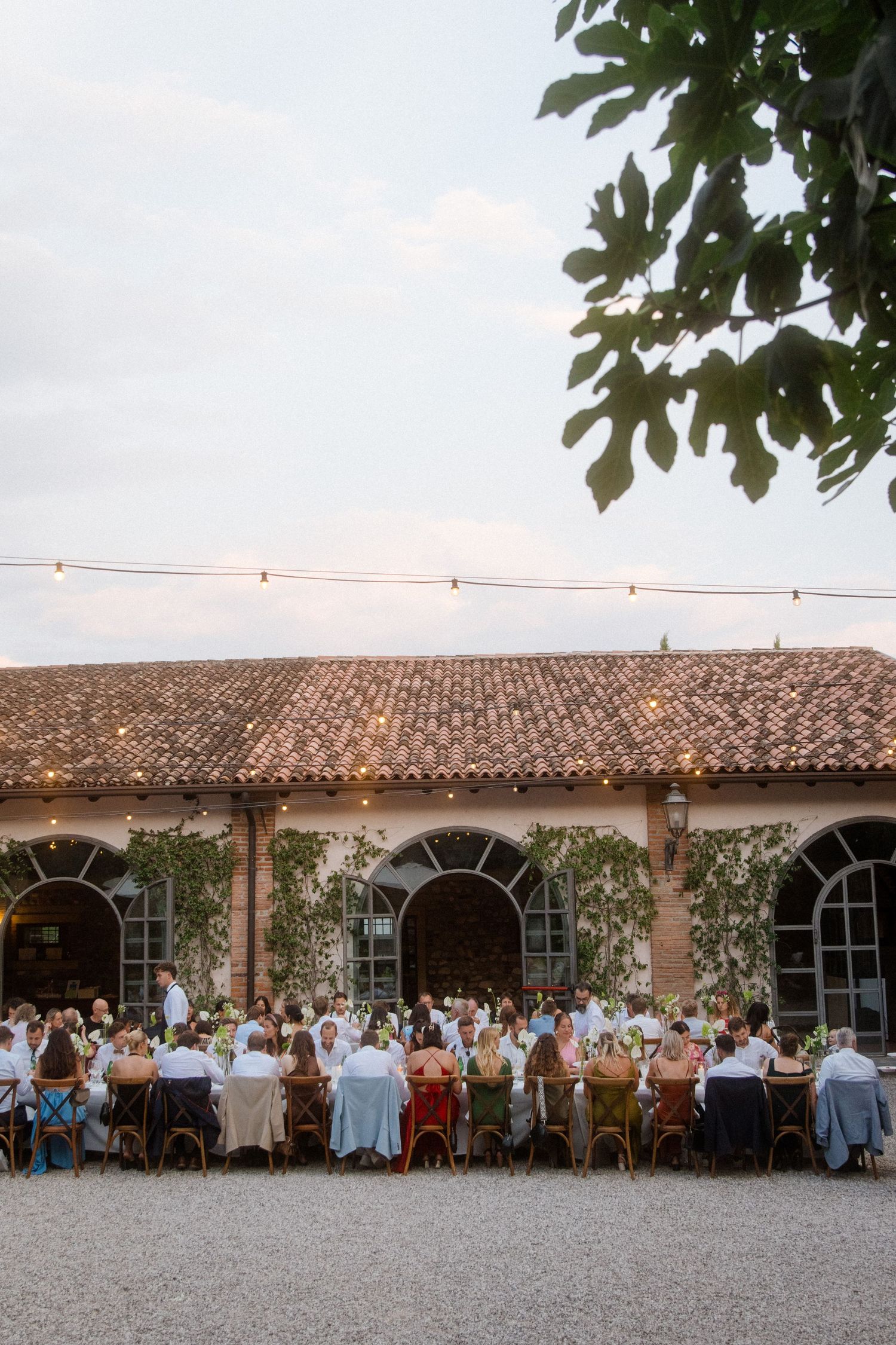 Outdoor wedding reception at a rustic venue with string lights and guests seated at long tables under a tiled roof.