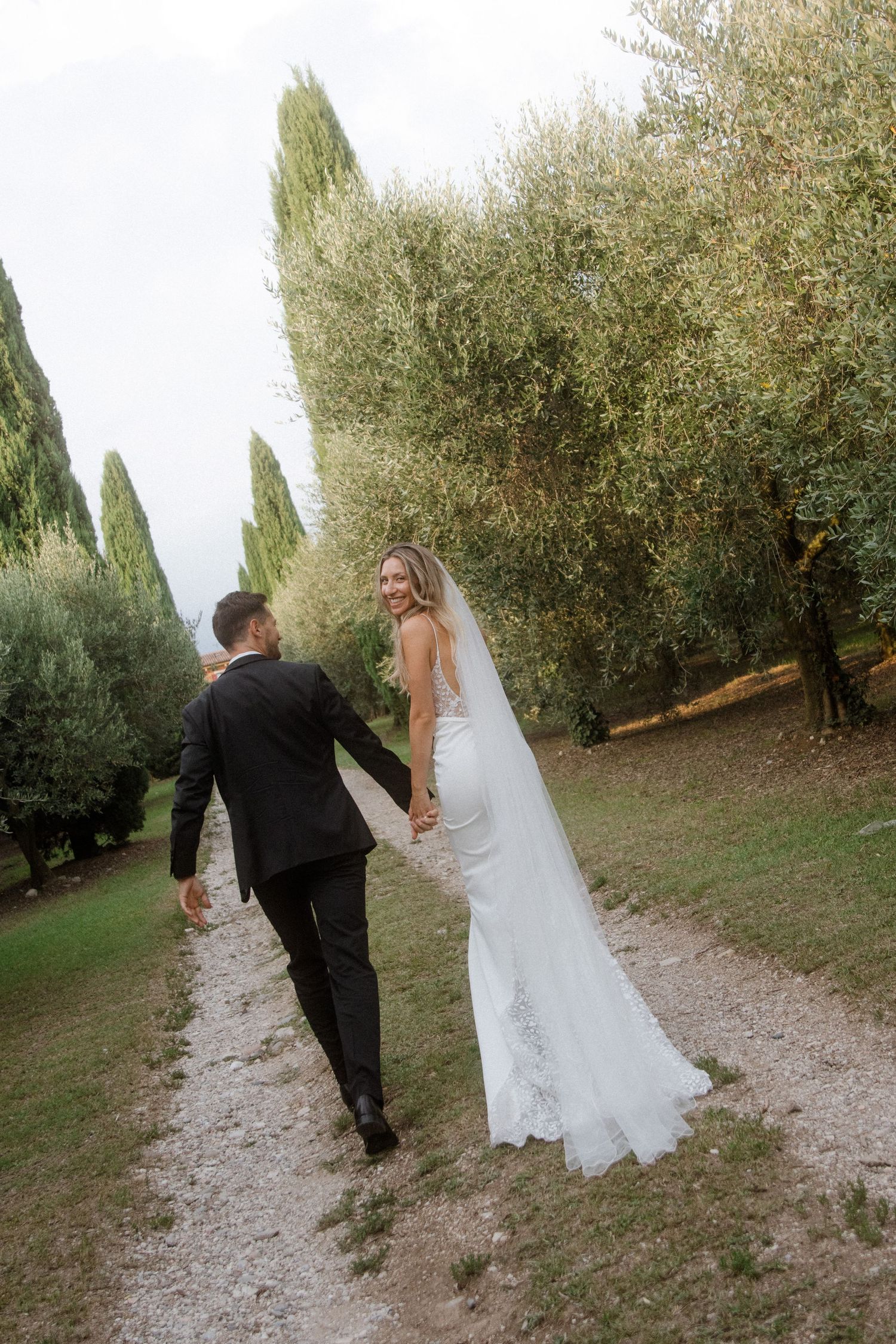 Couple walking hand in hand down a cypress-lined path in their wedding attire.