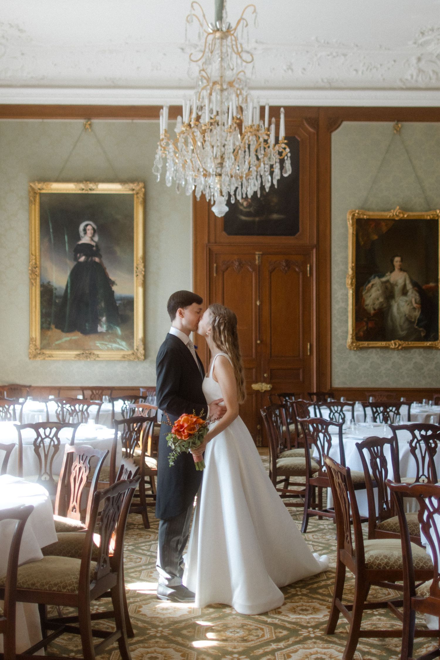 Elegant dining room with crystal chandeliers, ornate portraits, and traditional wooden chairs set for a formal event.
