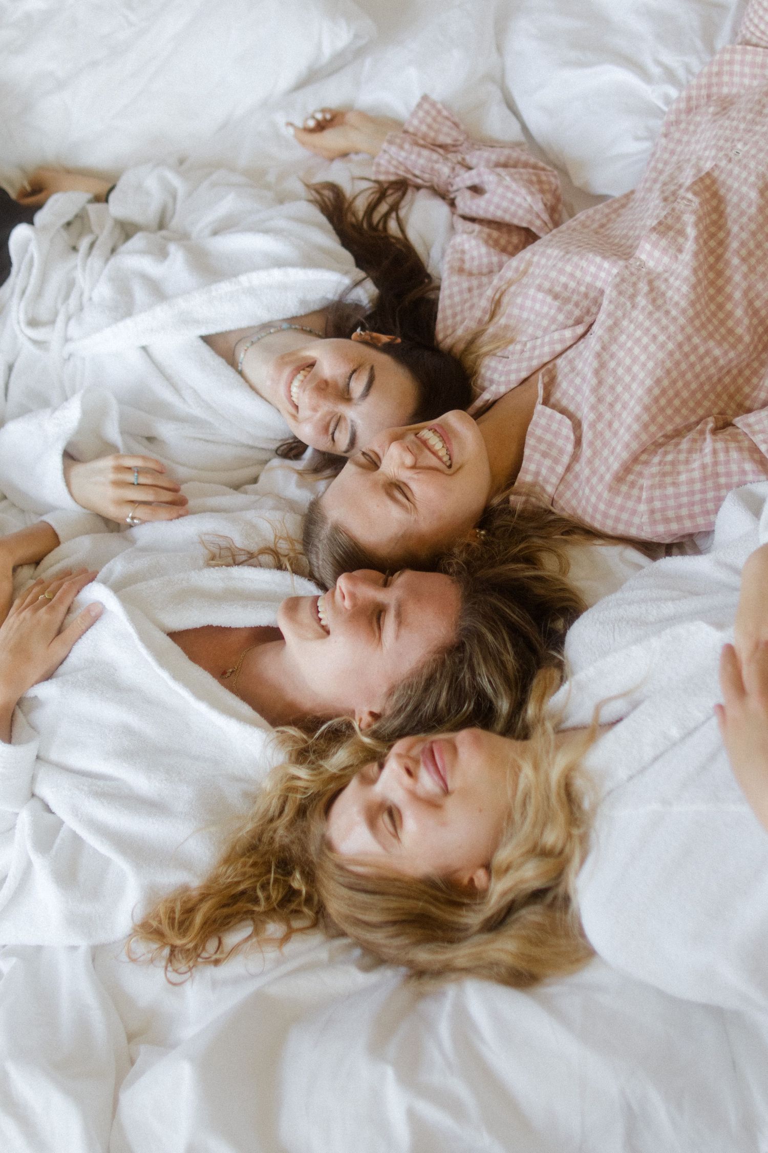 Women lying on white bedding share laughter and conversation together.