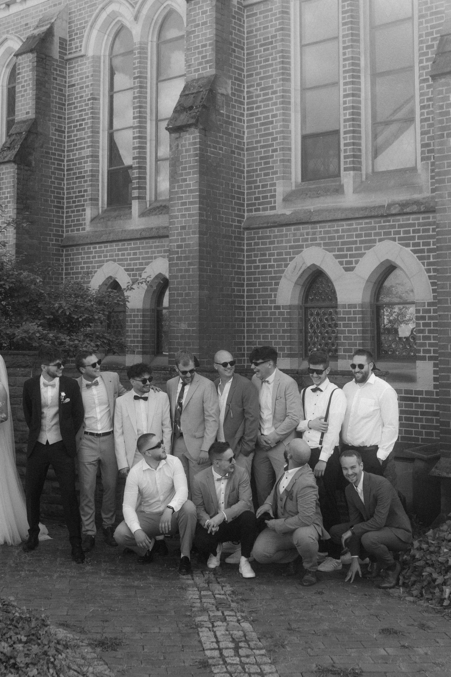 Black and white photo of groomsmen posing casually outside church building.