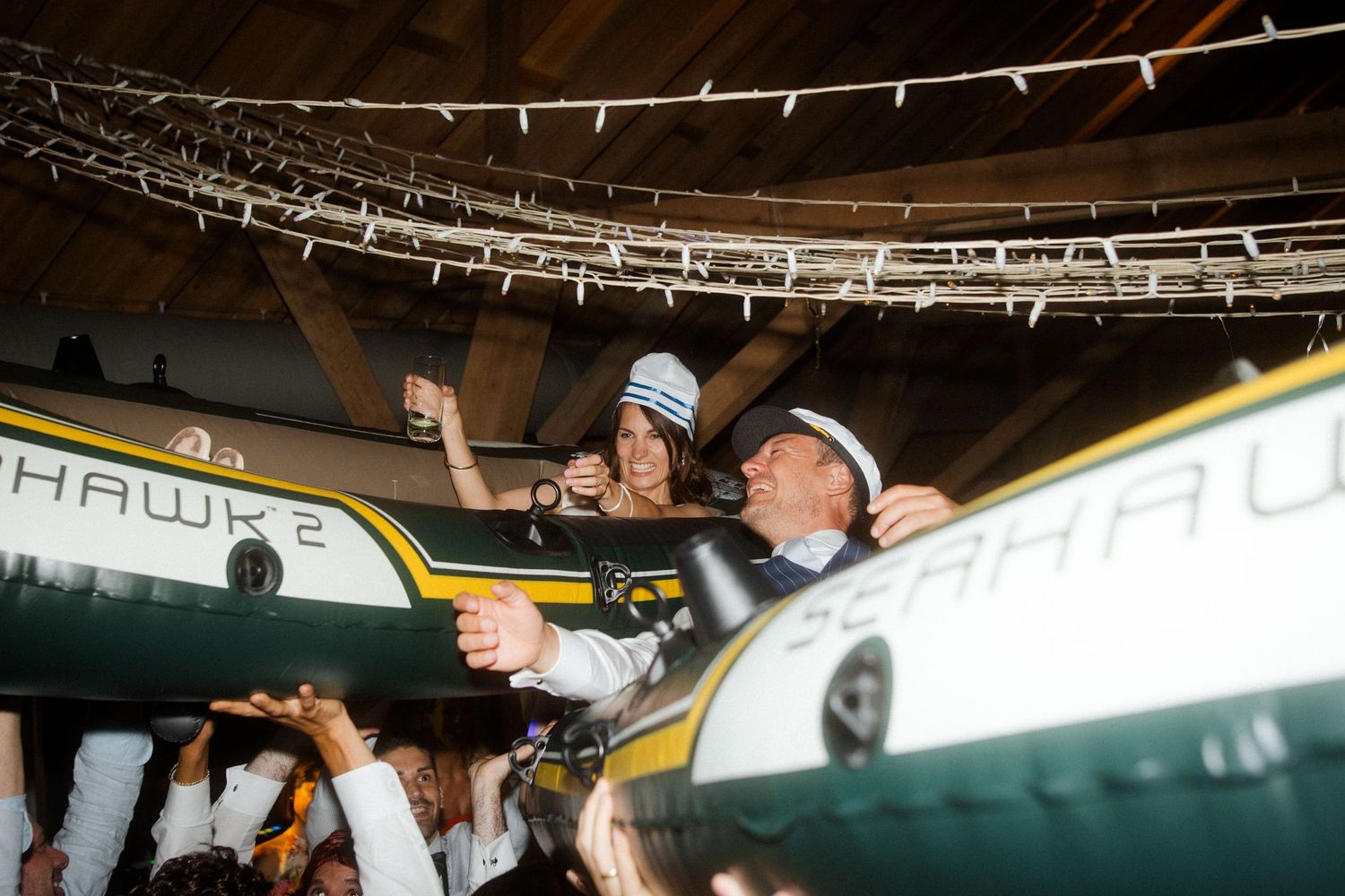 Partygoers lift person on inflatable boat during indoor celebration with string lights overhead.