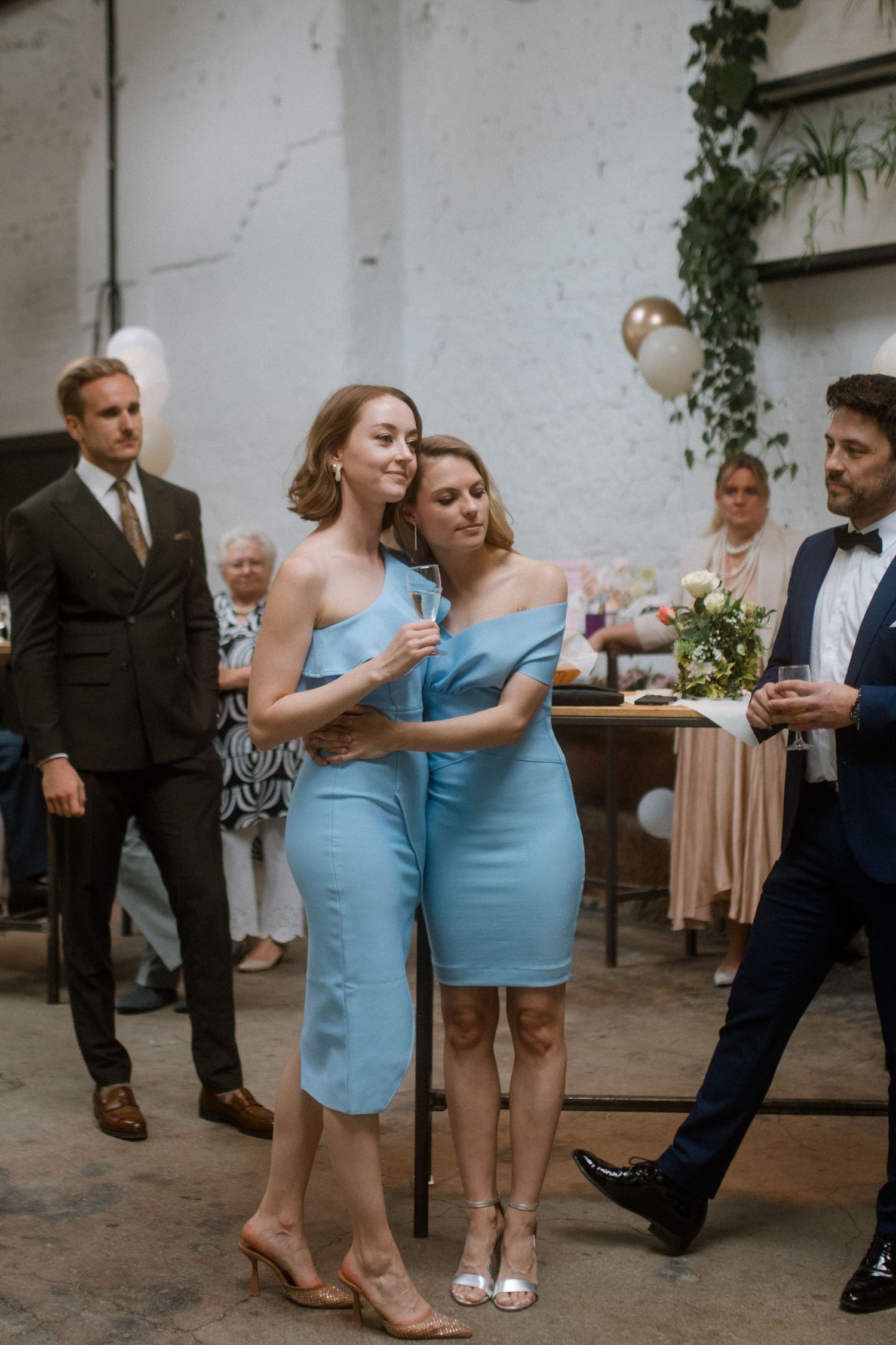 Wedding guests in blue dresses socialize during a reception in a white brick venue with greenery.