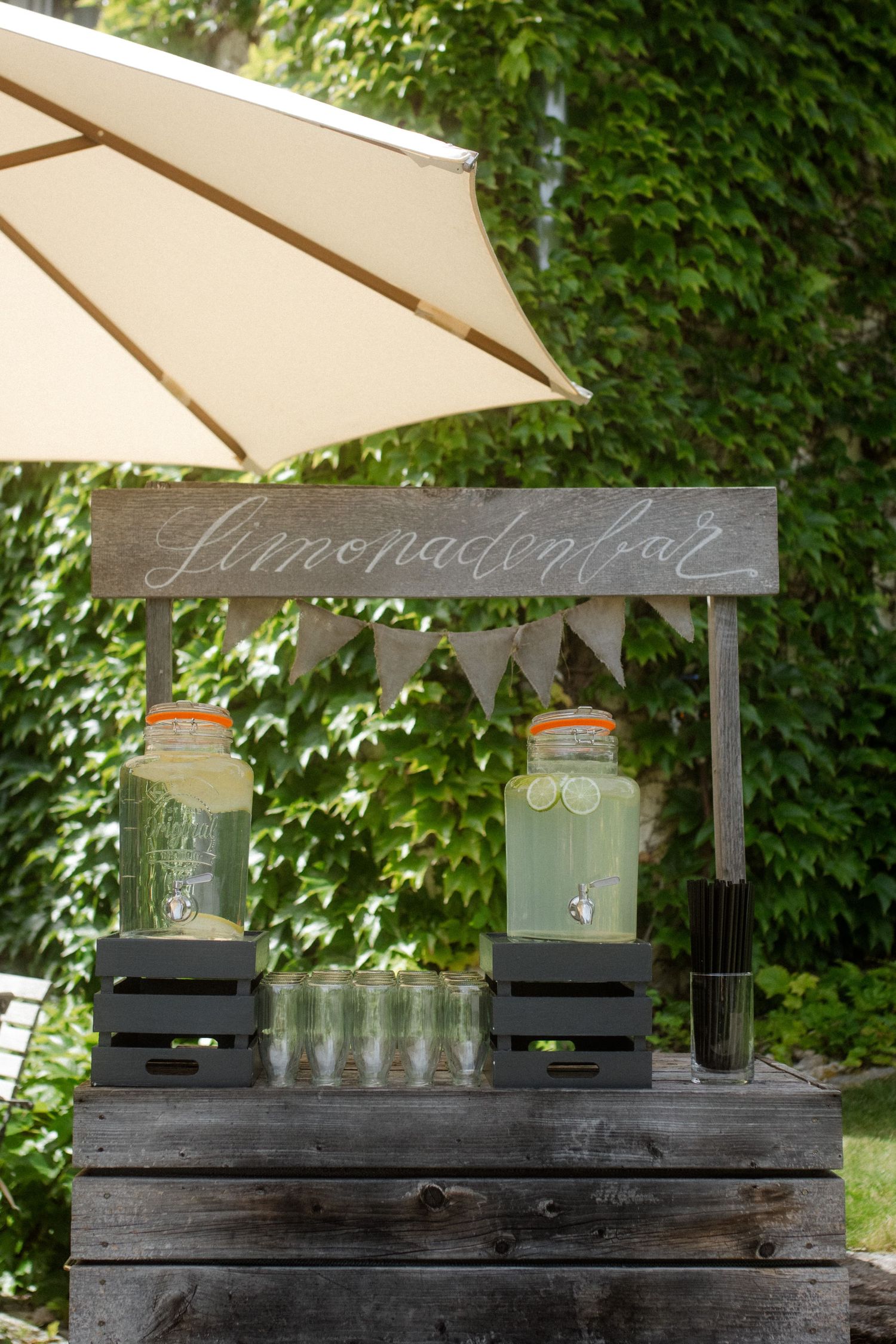 Rustic lemonade bar with glass dispensers under a cream umbrella and wooden sign.