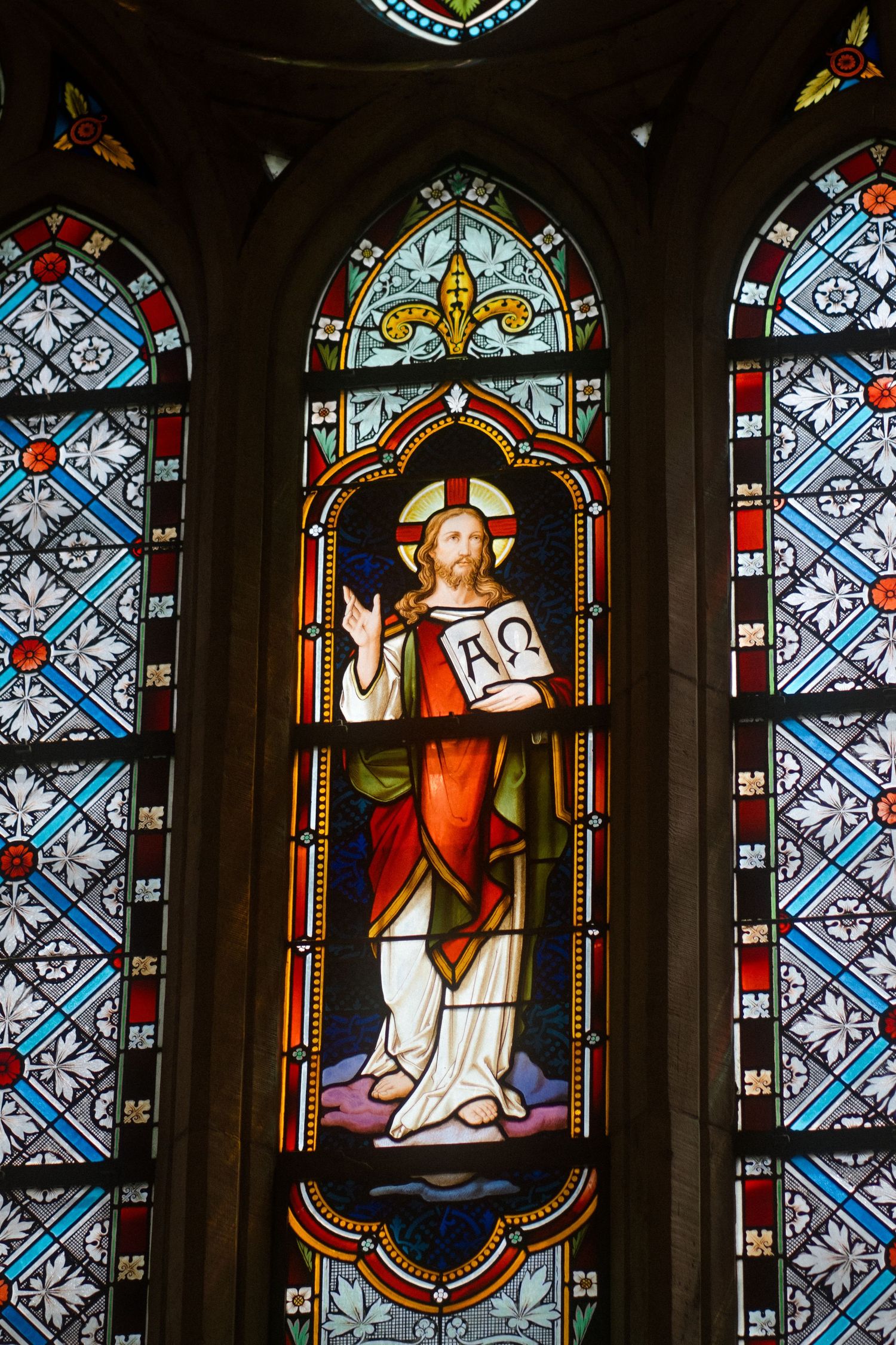 A stained glass church window depicting a religious figure holding a book with ornate Gothic-style patterns.