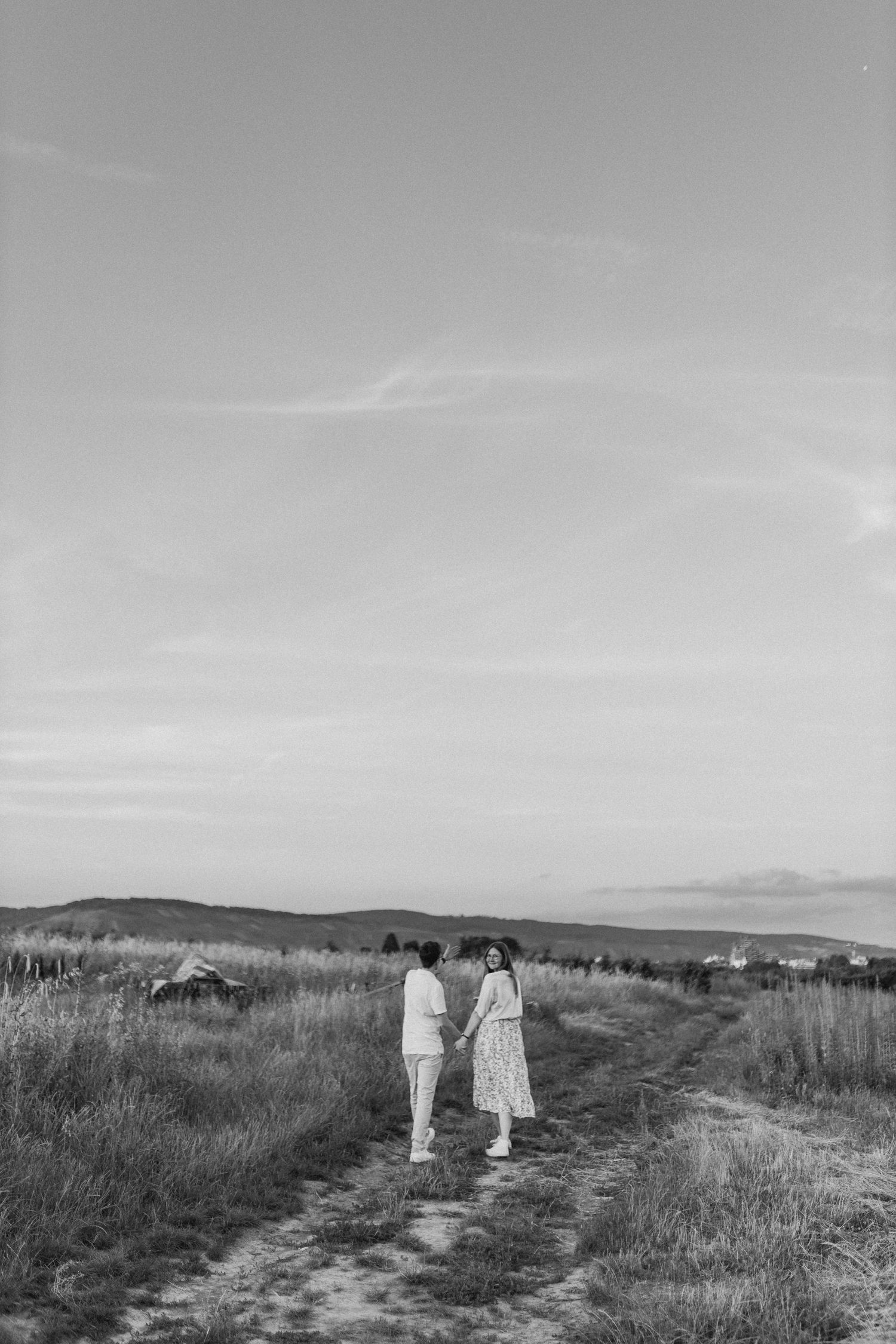A couple walks hand in hand through a serene field at sunset with mountains in the distance.