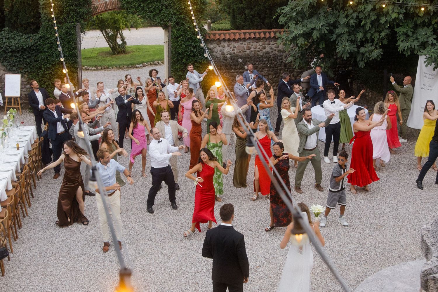 Wedding reception guests dance and celebrate outdoors on a gravel courtyard.