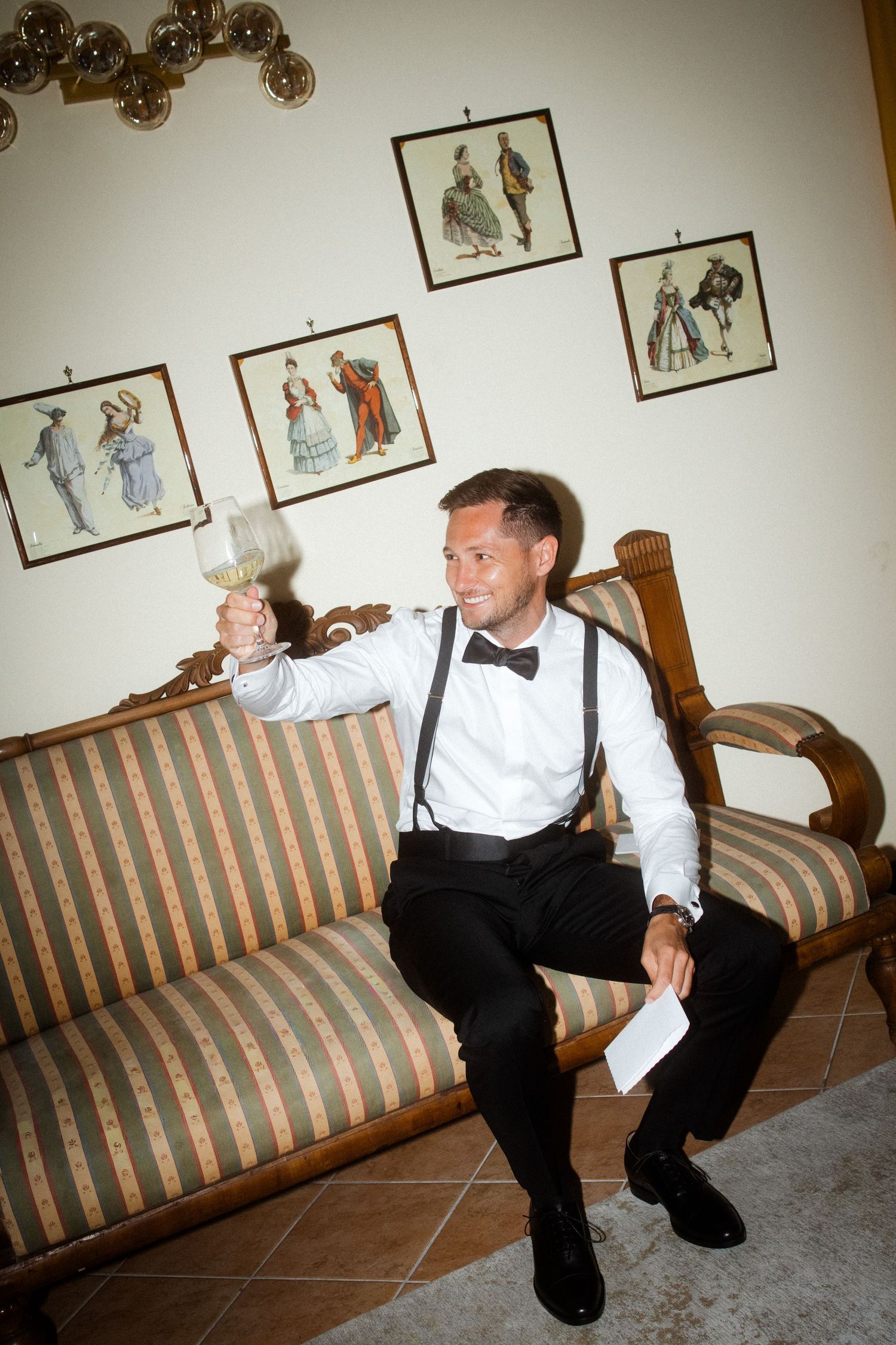 Someone in formal attire sits on a striped couch beneath framed artwork.