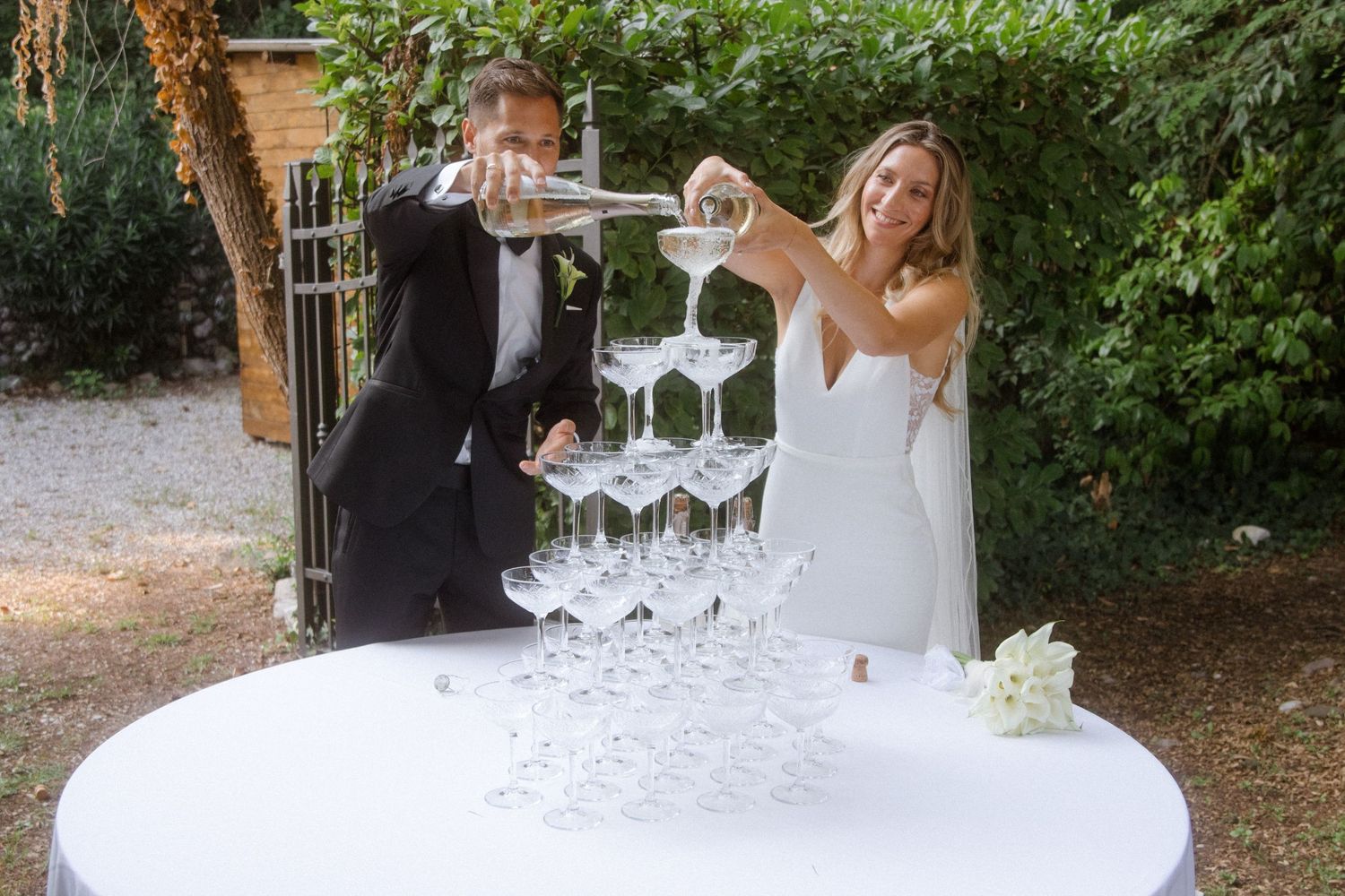 Wedding couple creating a champagne tower cascade in a scenic garden setting.