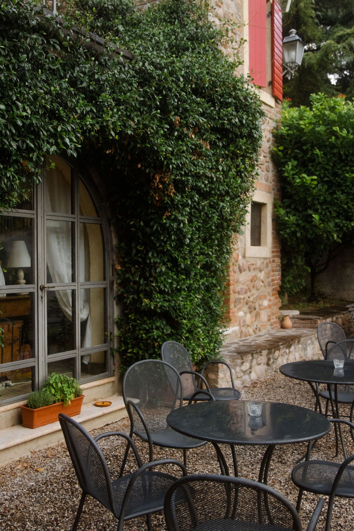 Rustic outdoor dining area with metal chairs against an ivy-covered stone building wall.