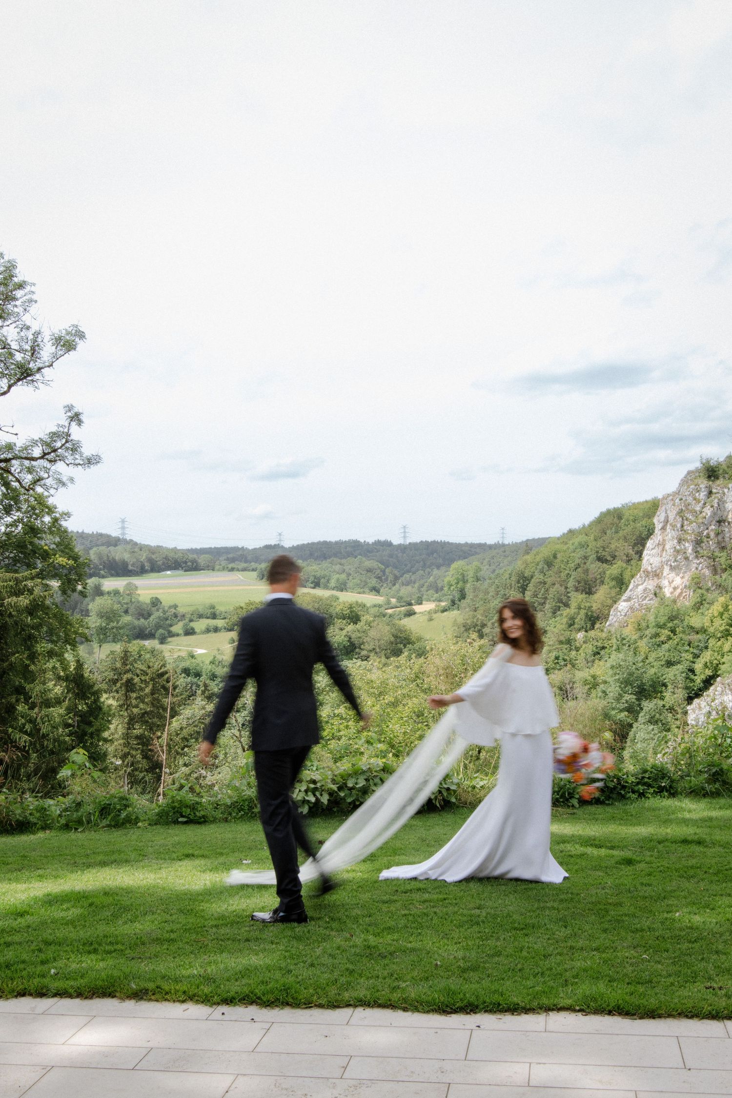 A wedding couple dances on a hilltop lawn overlooking a scenic valley with dramatic cliffs and green forests.