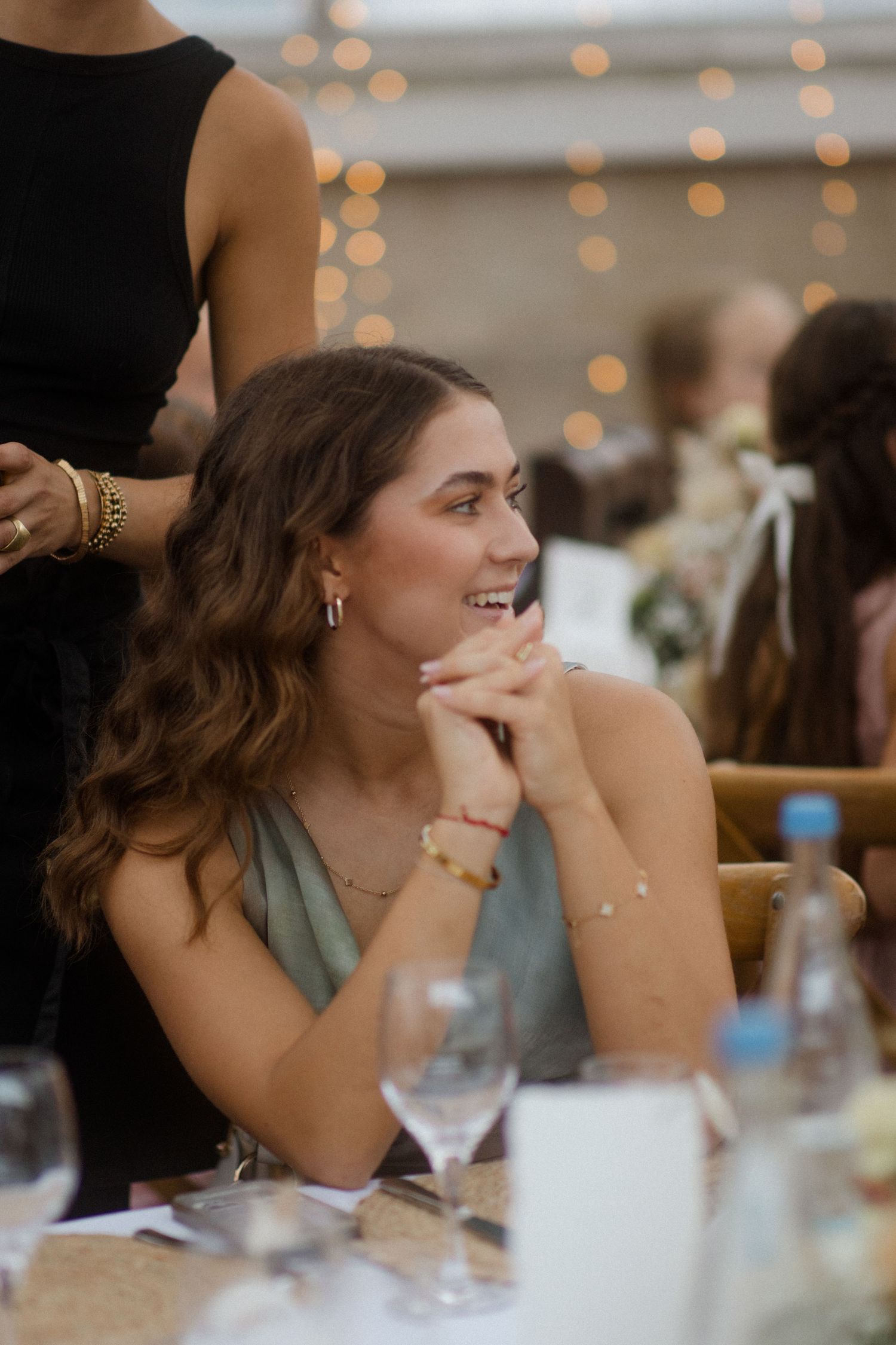 Guest at wedding reception smiling while seated at dinner table with string lights in background.