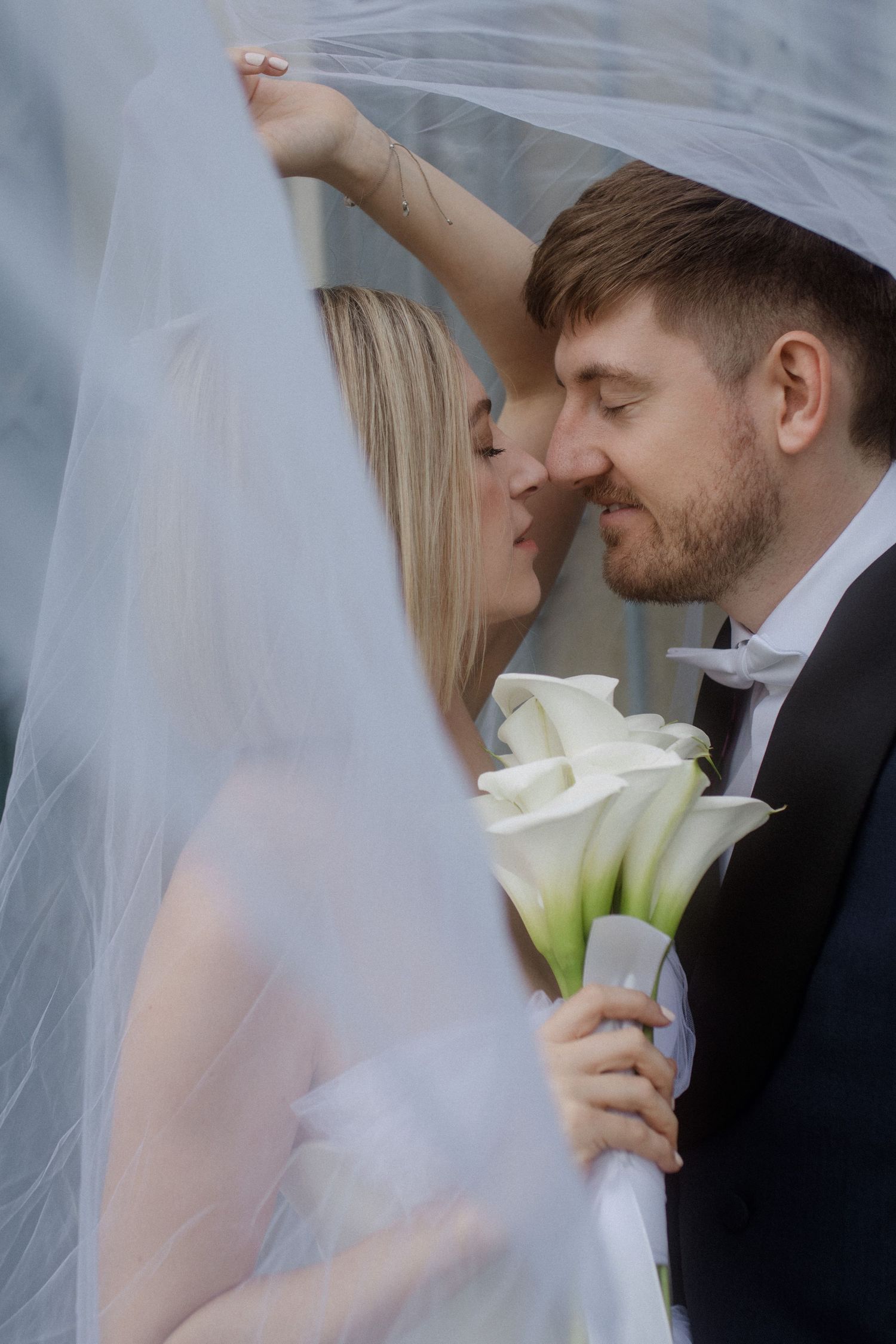 Tender moment between bride and groom under flowing veil with white calla lily bouquet.