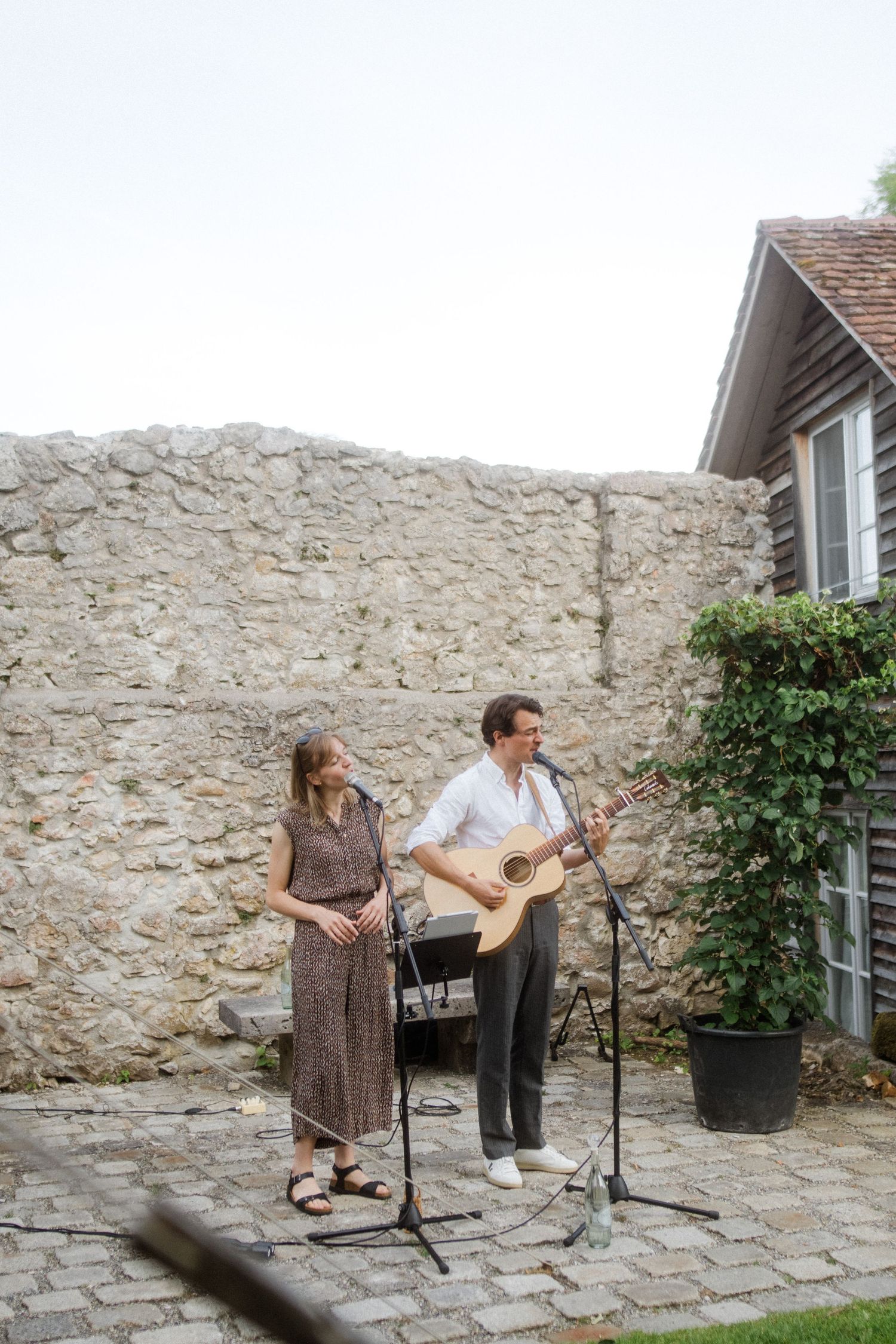 Musicians perform with guitar and microphones against a stone wall backdrop.