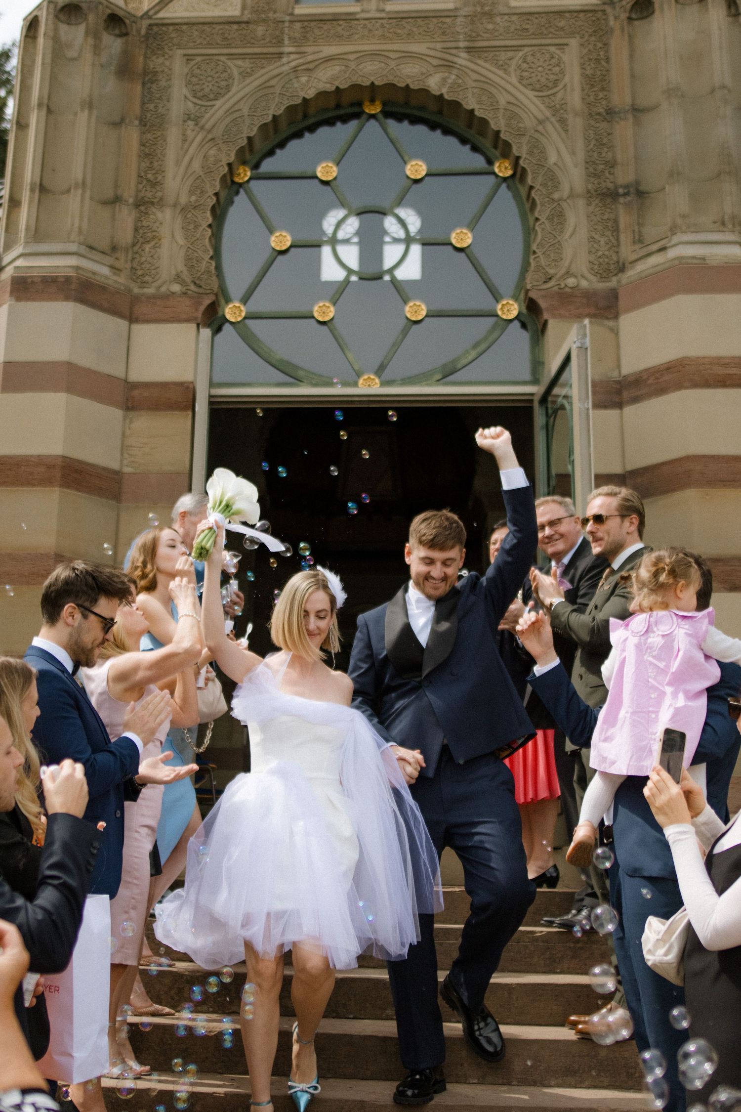 Newlyweds exiting church while guests blow bubbles under an ornate architectural doorway.