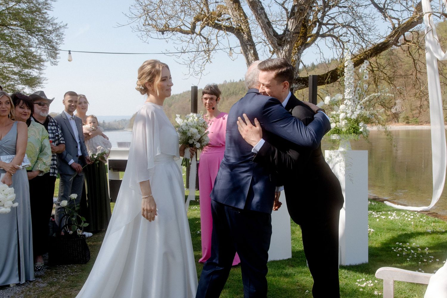 Bride in white gown embraces groom in navy suit at outdoor wedding ceremony by a lake.