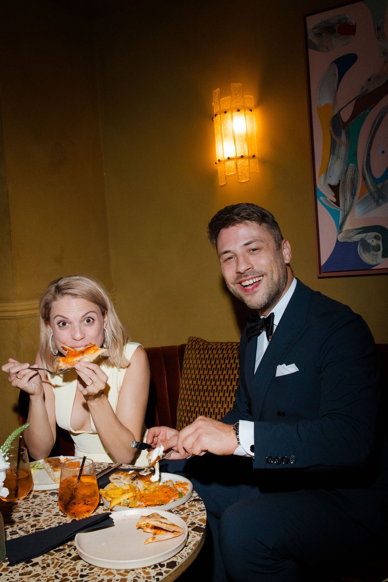 Elegant couple dining at a restaurant, woman eating pizza, man in tuxedo smiling, warm ambient lighting.