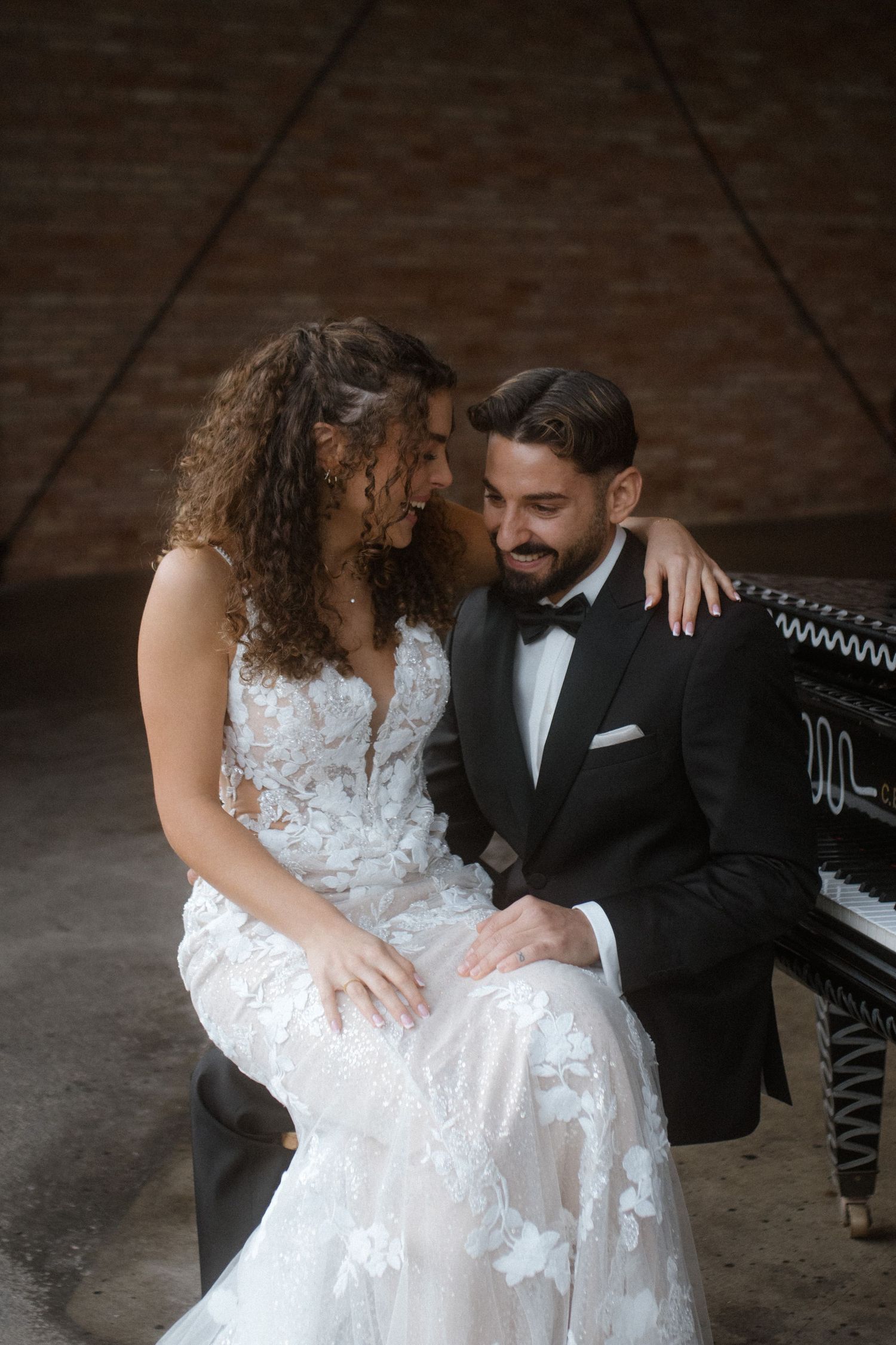 Intimate moment between couple at grand piano in moody brick-walled setting.