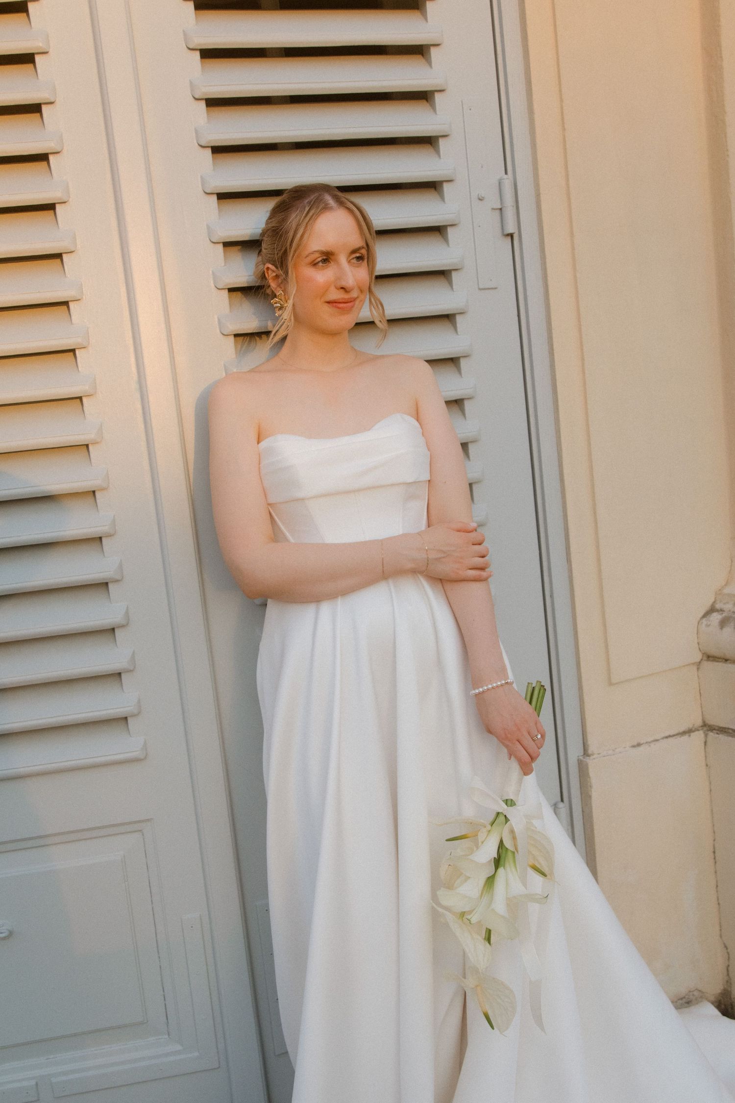 Elegant bride in flowing white dress holding calla lilies against shuttered doors.