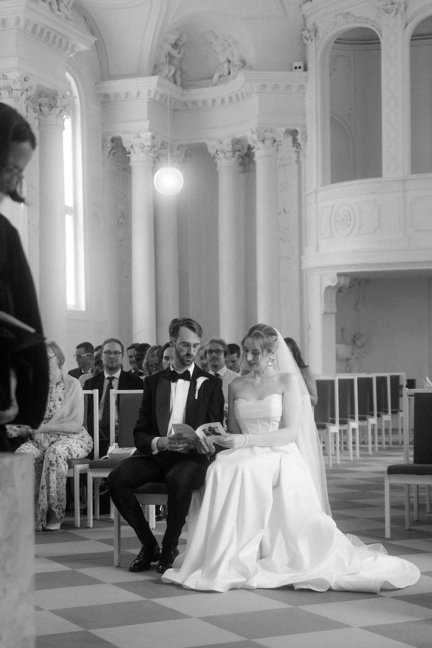 Black and white wedding photo in a historic hall with ornate columns and high ceilings.