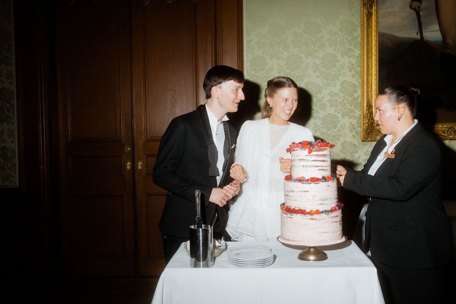Wedding party gathered around a three-tiered white cake with red decorations.