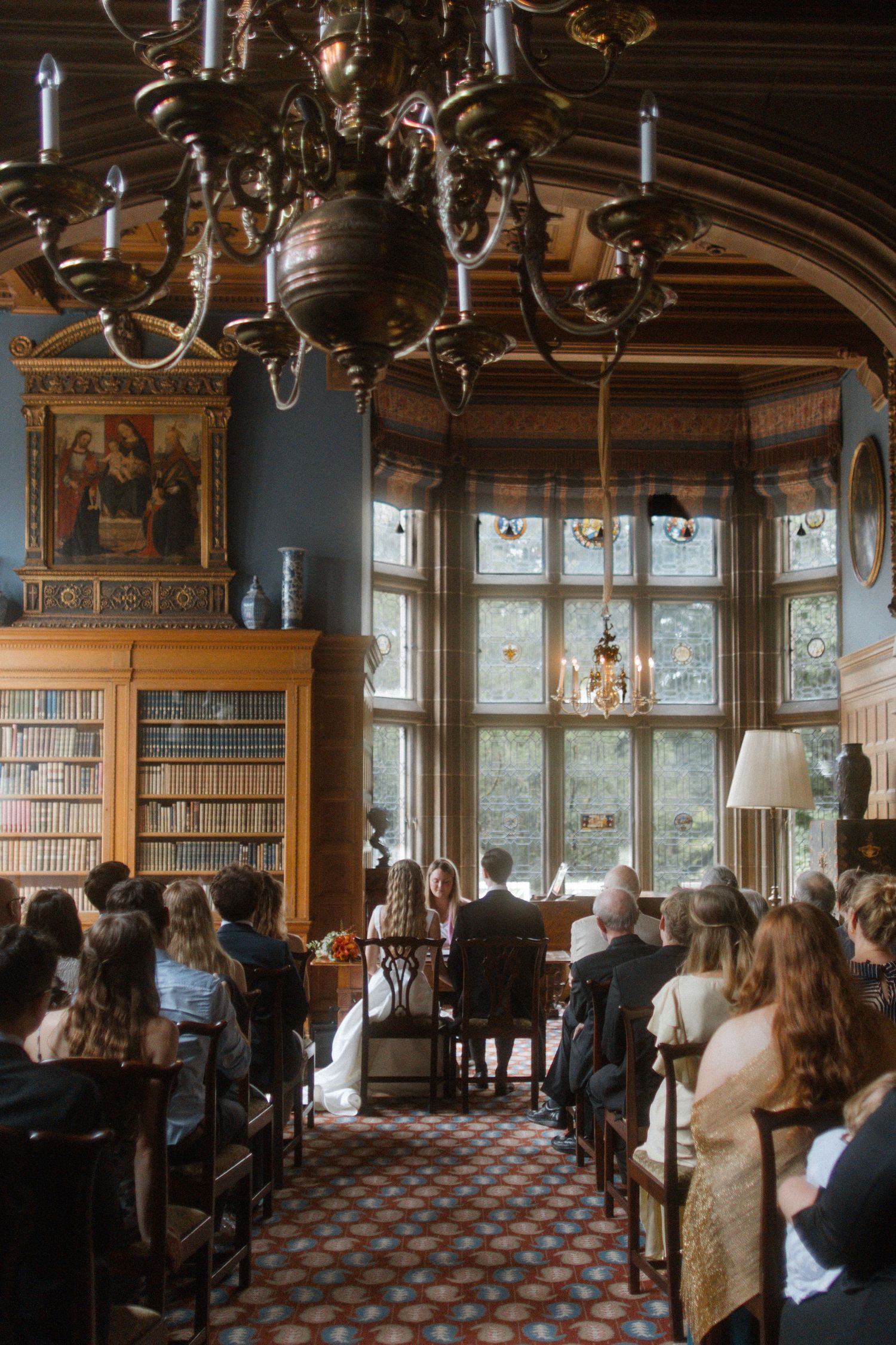 Wedding guests gathered in a historic library with chandeliers and large windows.