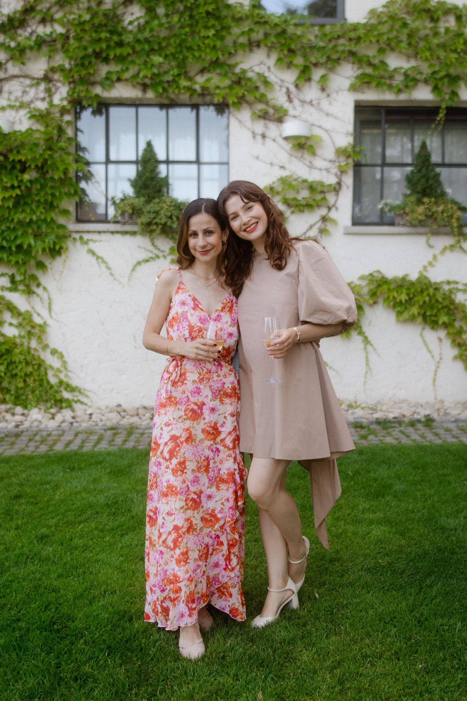 Two guests in floral and beige dresses pose against an ivy-covered white building.