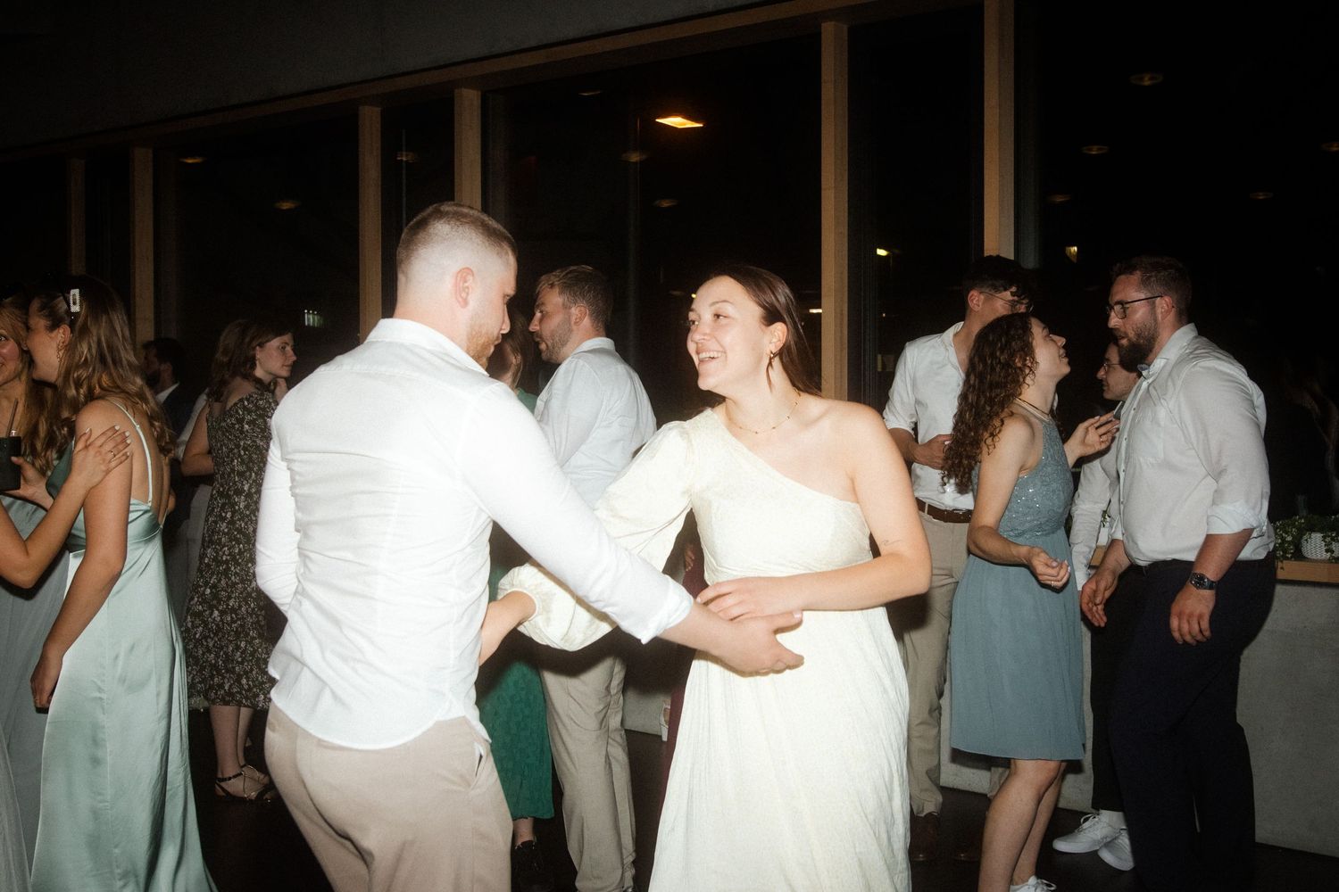 Wedding guests dancing together during reception with lights reflecting in windows.